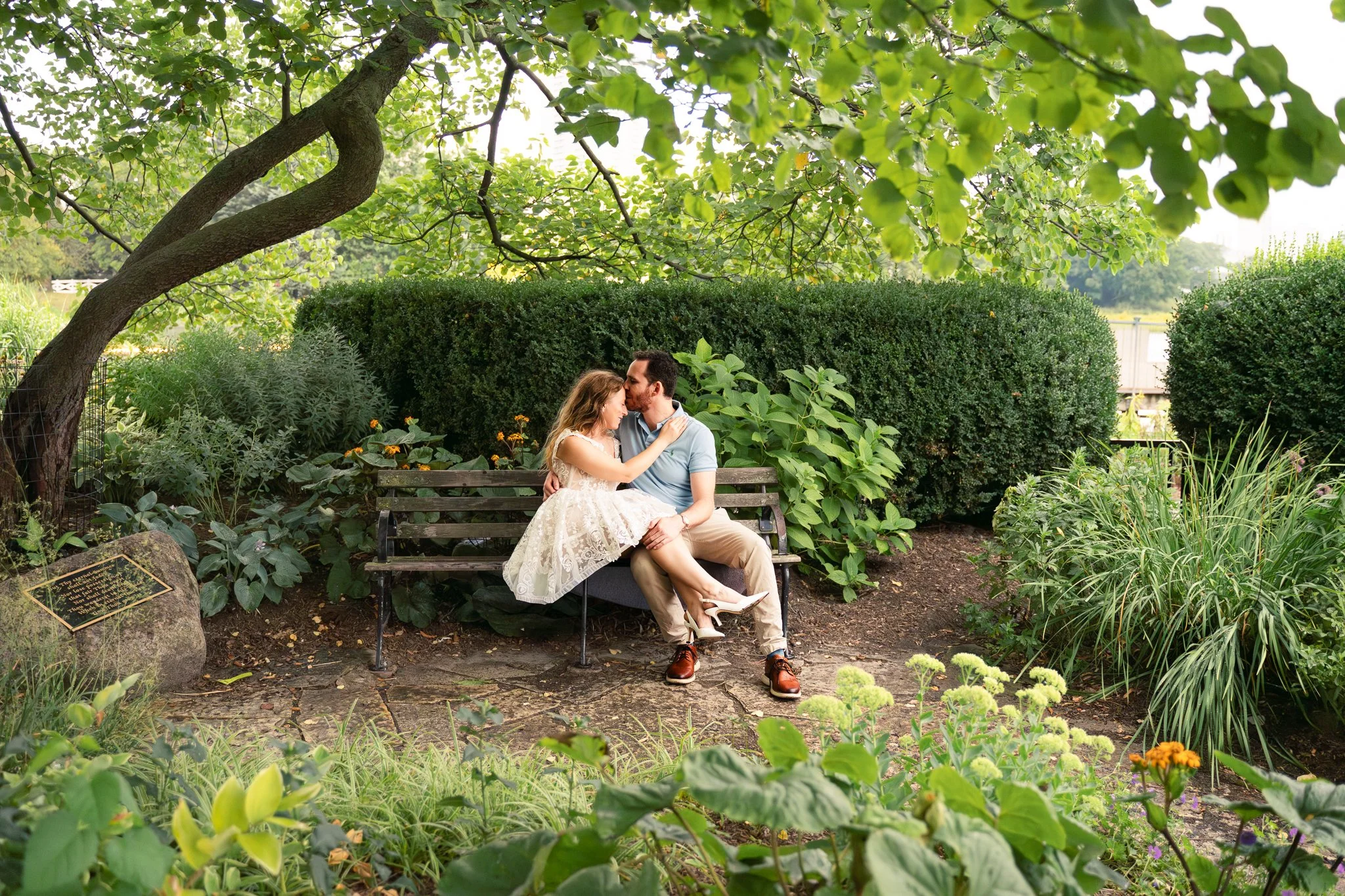 Lincoln Park Chicago Illinois Engagement Photographer | Bride in lace white dress sitting with legs across groom on park bench in garden, groom kisses her forehead | Midwest Wedding and Engagement Photography Sessions