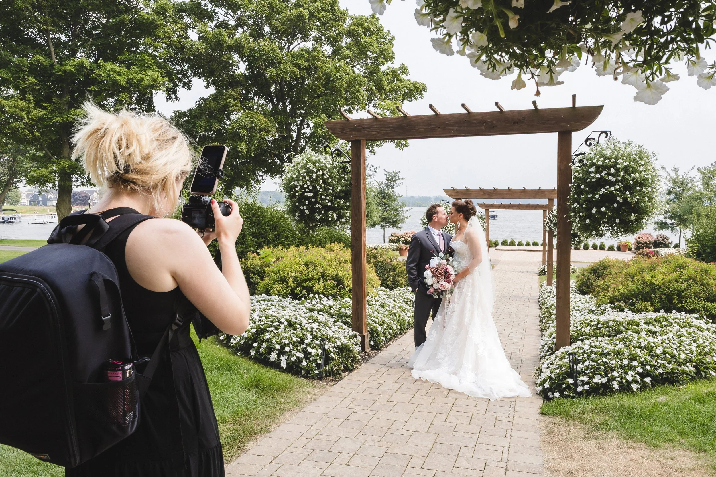 A bride and groom kiss under a wooden arch on a pathway by a river, with a female photographer taking a picture of the scene.