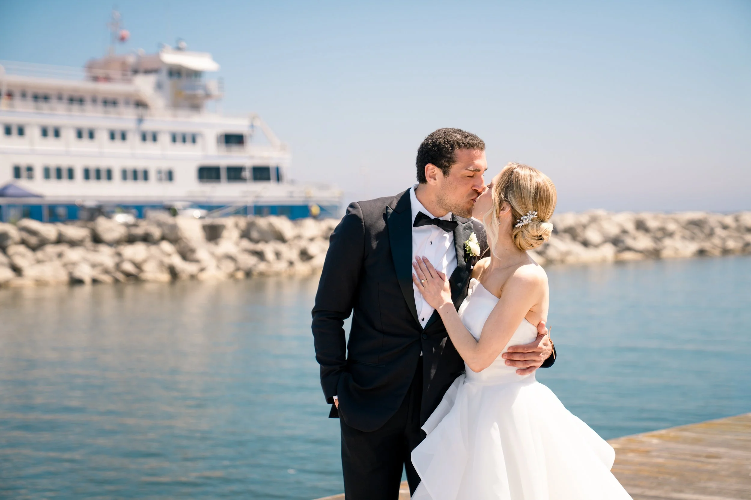 A bride and groom share a kiss near a waterfront, with a large cruise ship docked in the background on a sunny day.