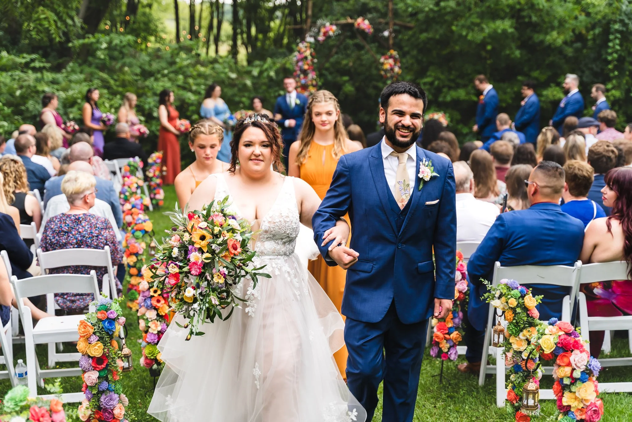 Bride and groom walking down the aisle at a wedding ceremony outdoors, surrounded by guests and colorful floral decorations.