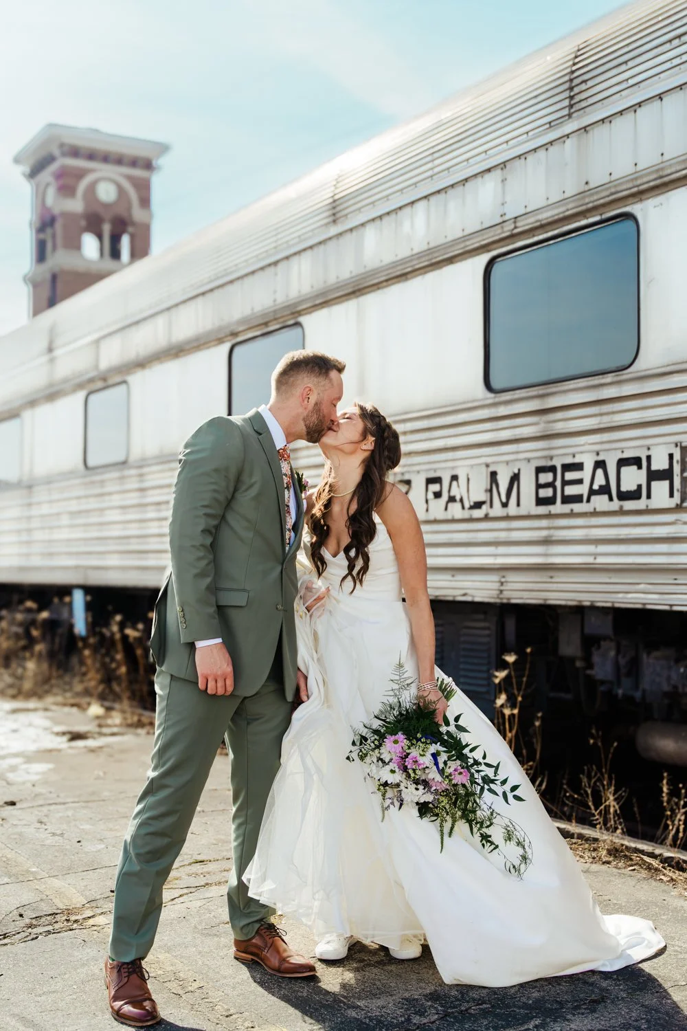 A bride and groom sharing a kiss in front of a vintage train labeled 'Palm Beach.' The groom wears a gray suit and brown shoes, and the bride is in a white wedding dress holding a bouquet of flowers.