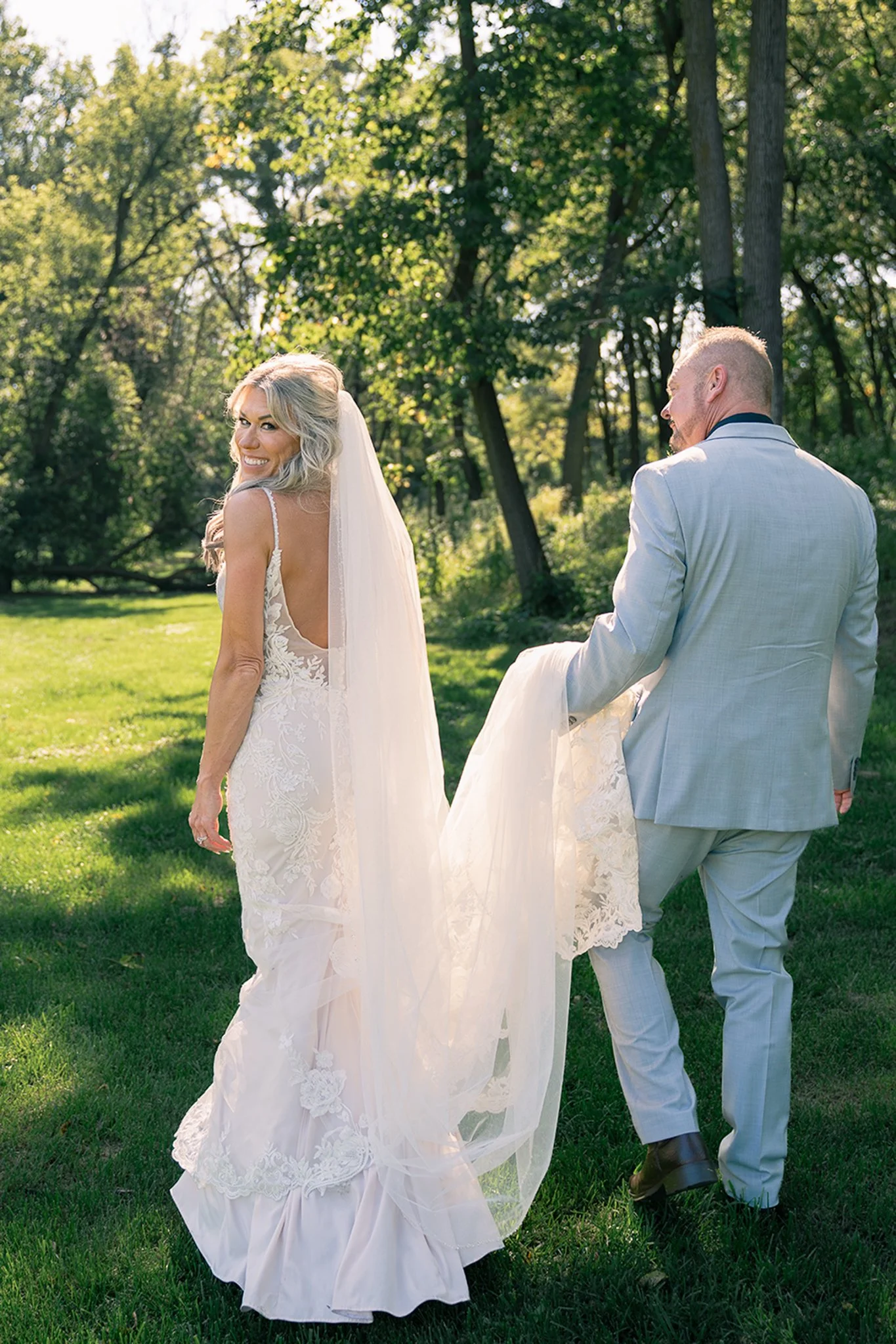 Best rated Wisconsin Wedding Photographer | Bride and groom portraits, groom carries the train of bride's long lace wedding dress, bride smiles and looks back at camera