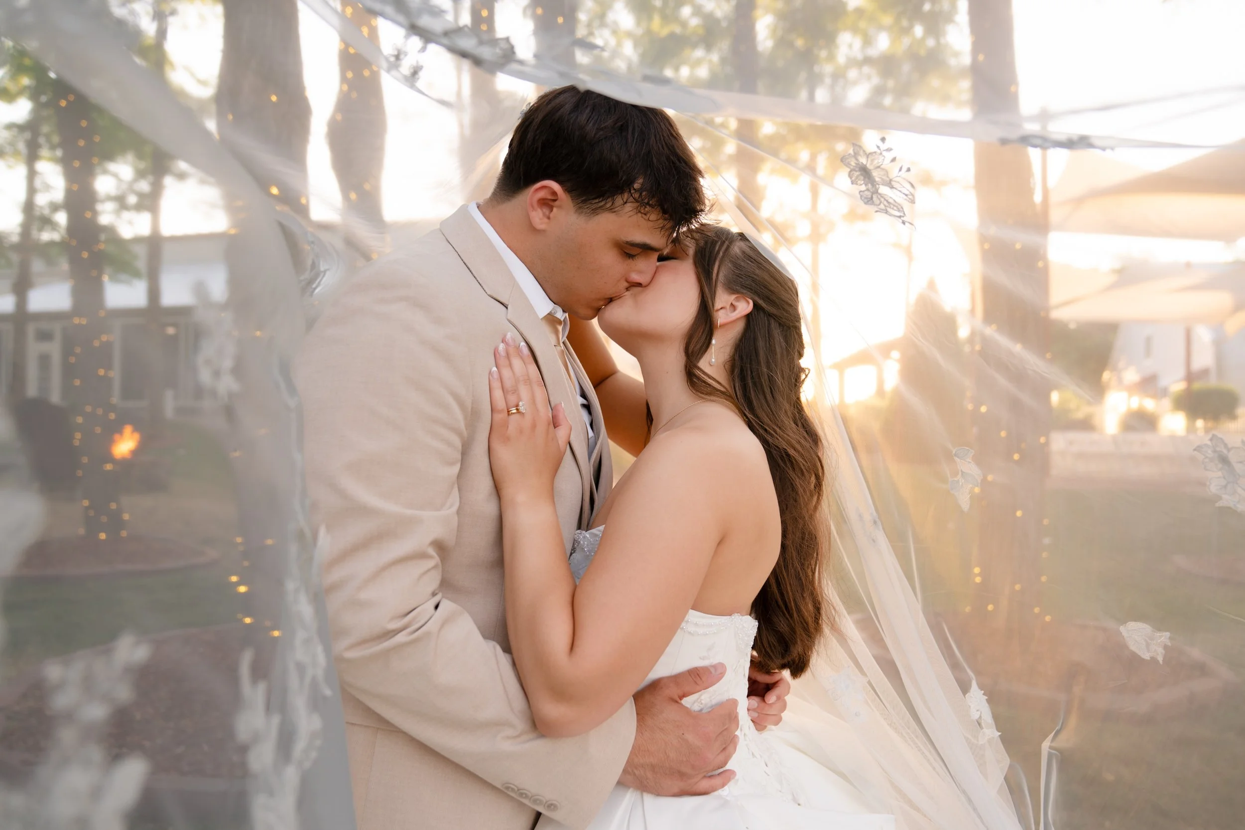 A newlywed couple sharing a kiss under a decorated tent at sunset, outdoors in a park or garden setting.