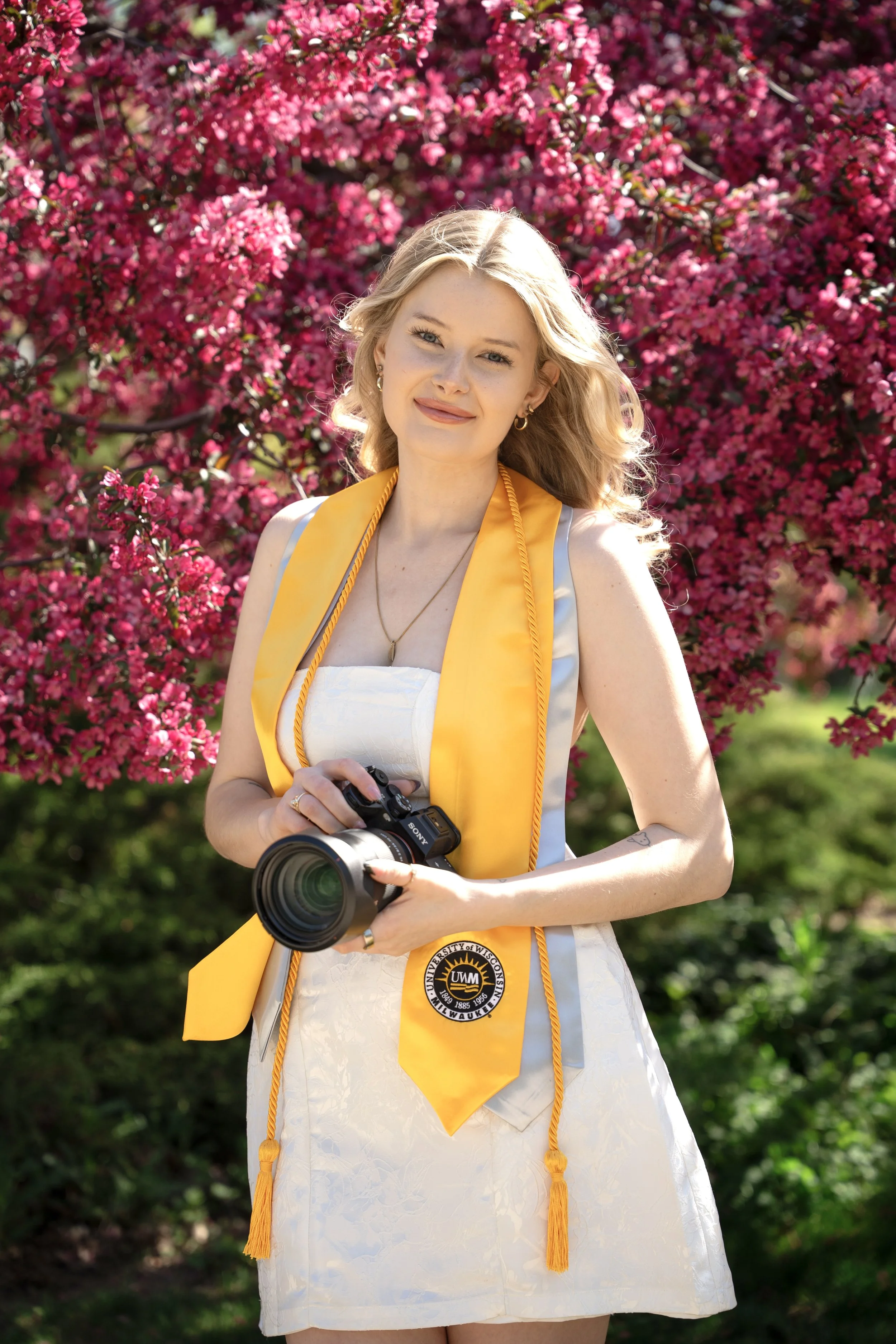A young woman in a white dress and a yellow graduation stole with the University of Wisconsin-Milwaukee emblem, holding a camera, standing in front of pink flowering bushes.