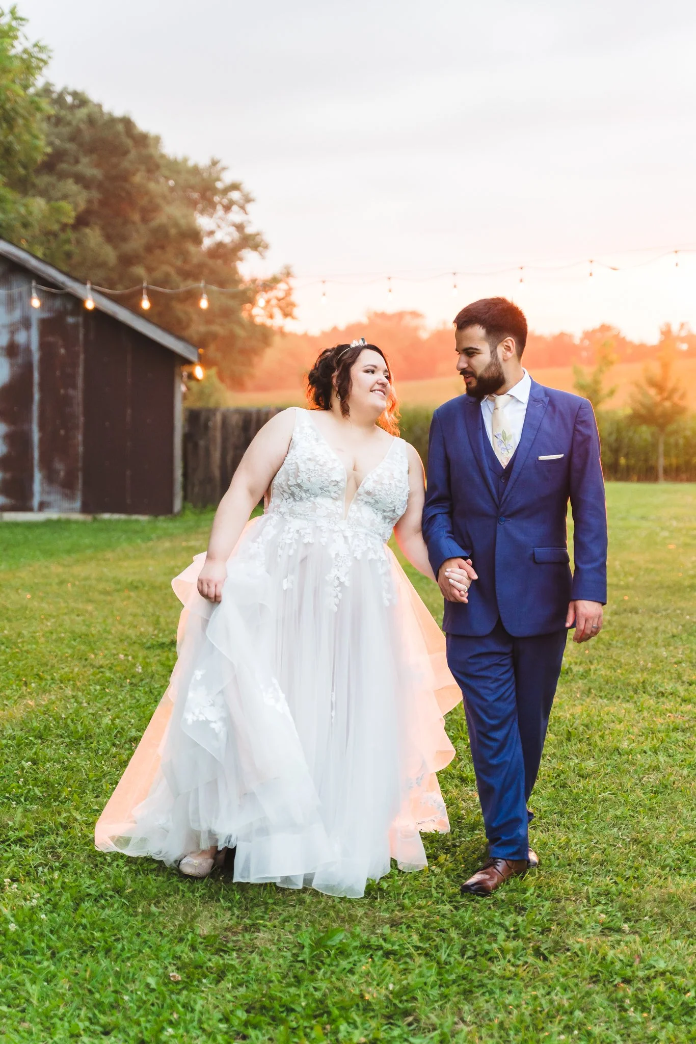 A newlywed couple walking hand-in-hand on a grassy field at sunset. The bride is wearing a white wedding dress with lace details, and the groom is dressed in a blue suit with a white shirt and tie.