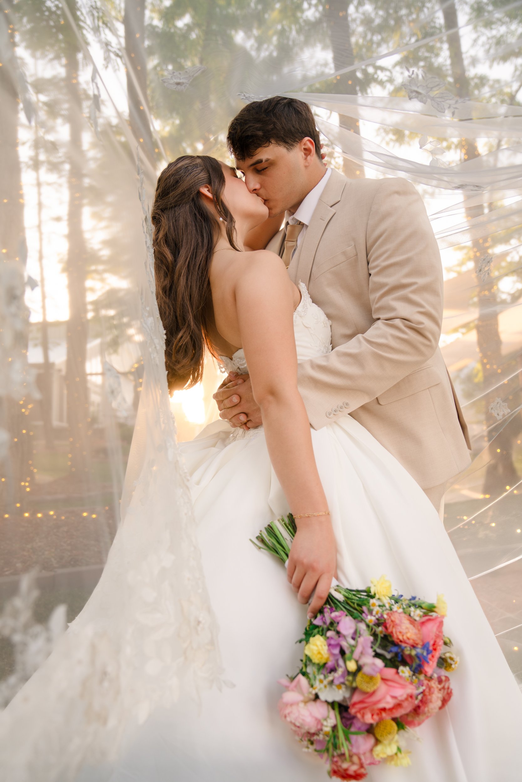 A bride and groom sharing a kiss during their wedding ceremony. The bride is holding a bouquet of colorful flowers, and they are standing under a canopy with fairy lights and trees in the background.