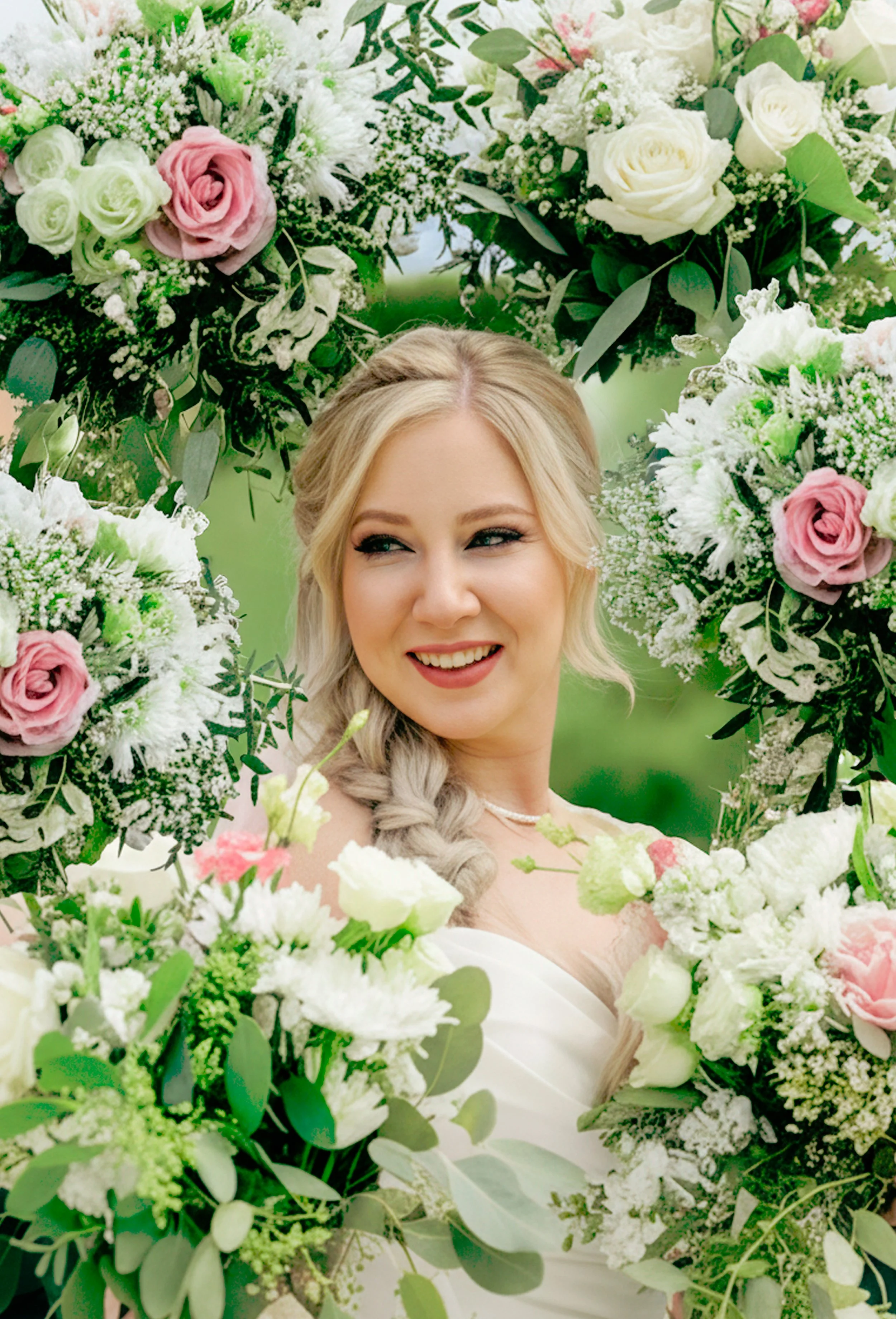 A smiling woman with blonde hair styled in a braid, surrounded by white, pink, and green flowers in a floral wreath, outdoors with greenery in the background.