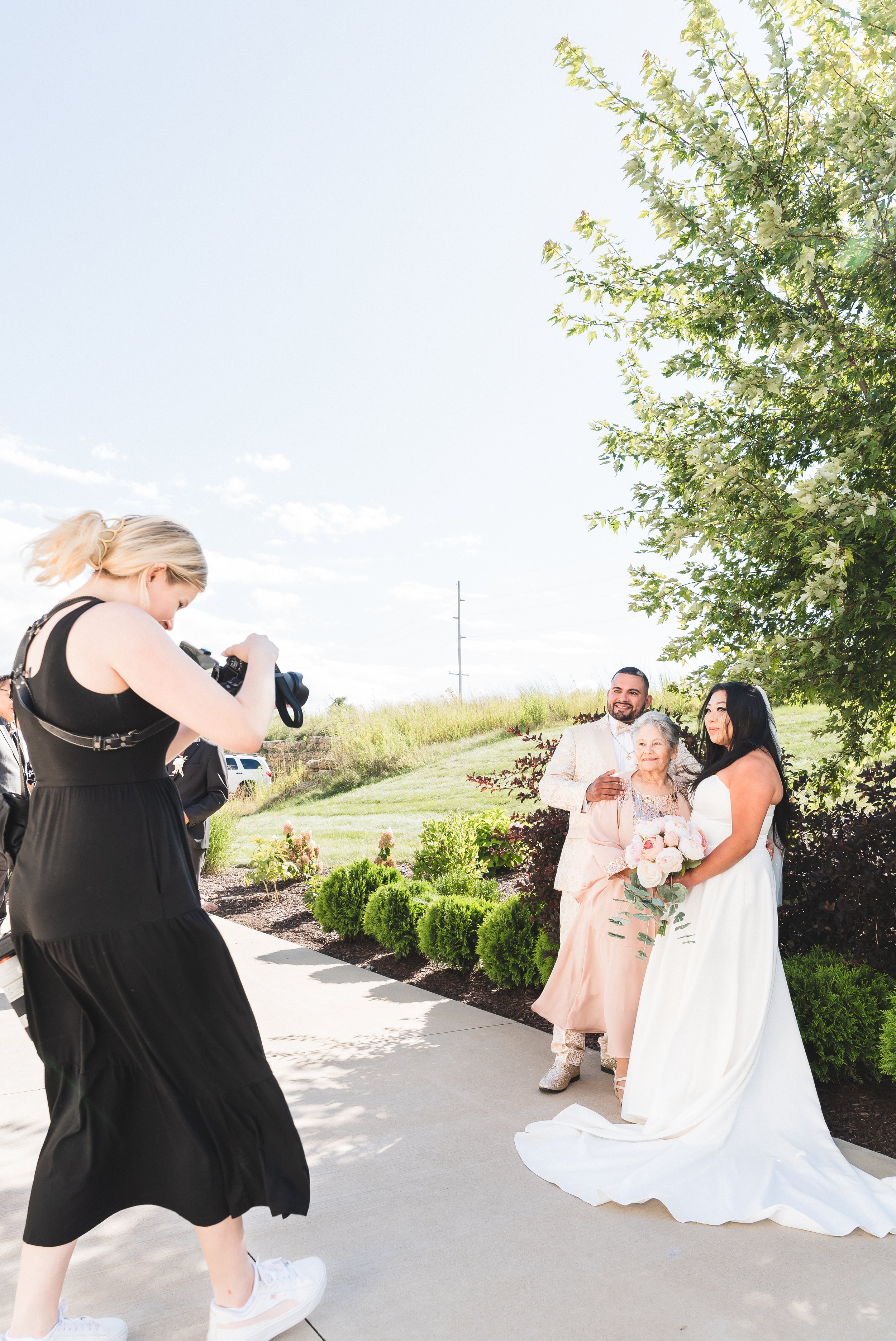 A bride in a white wedding gown holding a bouquet of pink and white flowers, standing next to an elderly woman in a pink dress and a man in a beige suit, all outdoors under a tree, being photographed by a woman in a black dress
