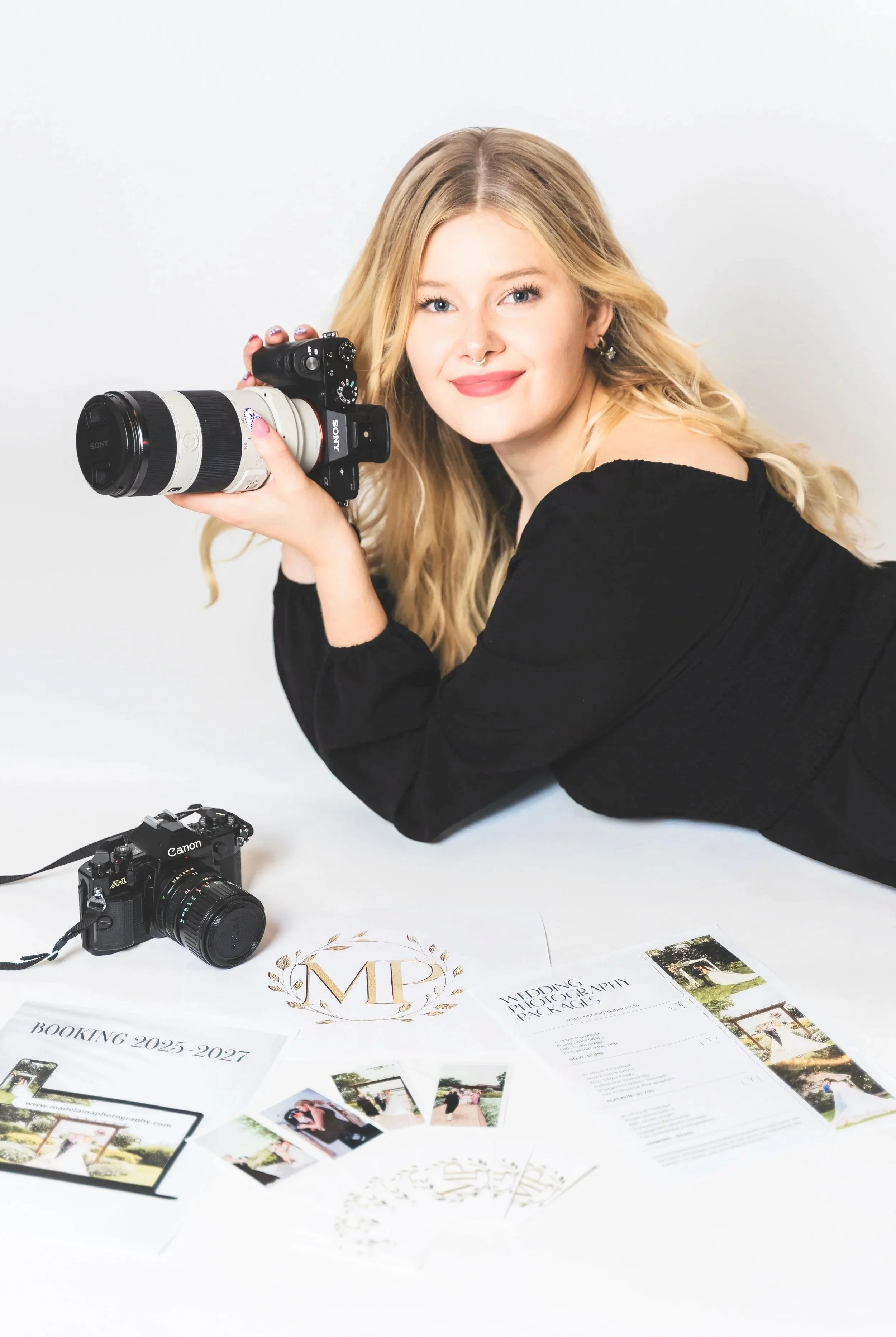 A woman with blonde hair, dressed in black, holding a camera with a large lens, looking at the camera and smiling. There are two cameras on a white table with promotional materials and photos for a photography business.