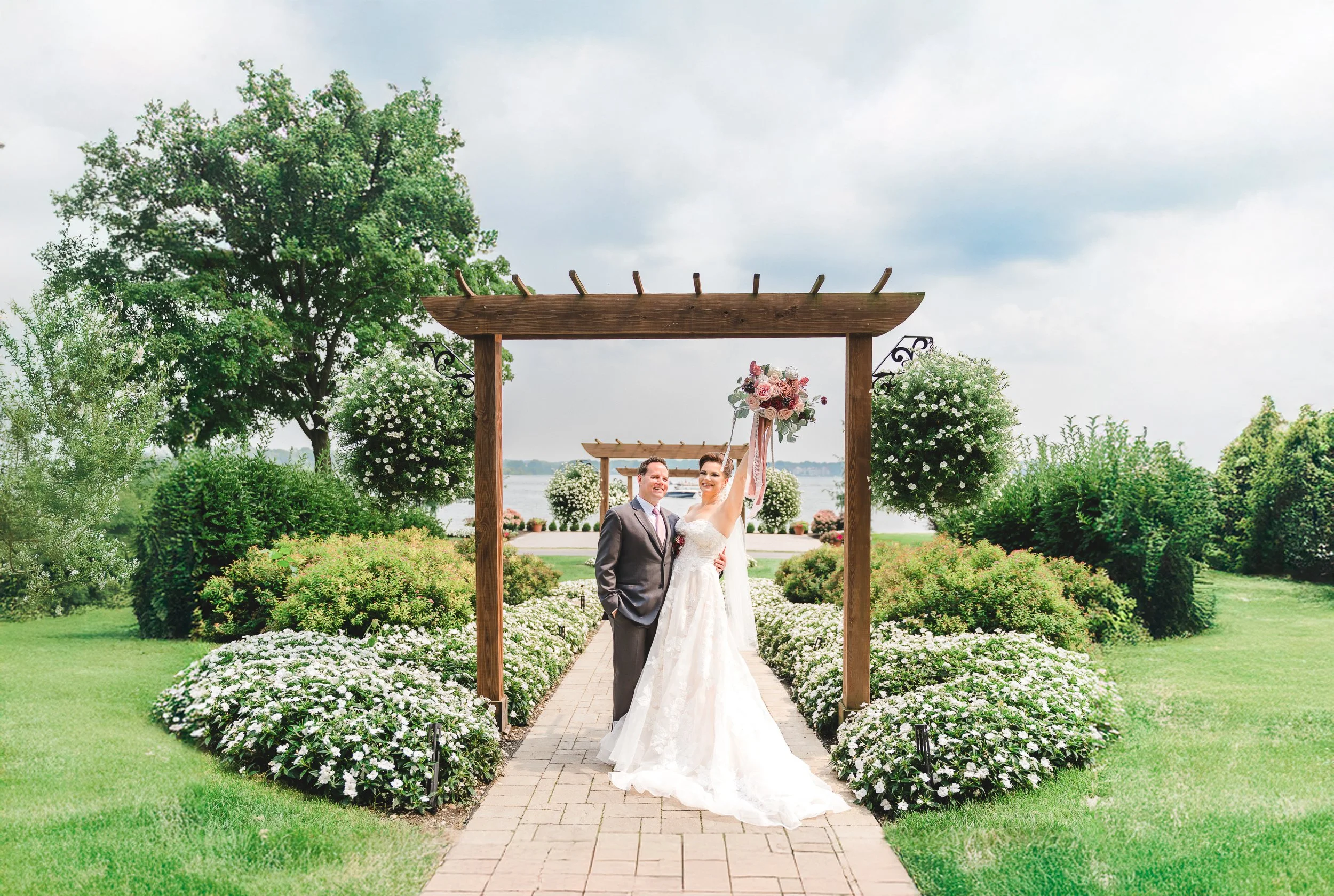 A bride in a white wedding gown and a groom in a gray suit standing under a wooden wedding arch on a garden path, holding a bouquet of flowers, with greenery and a lake in the background.