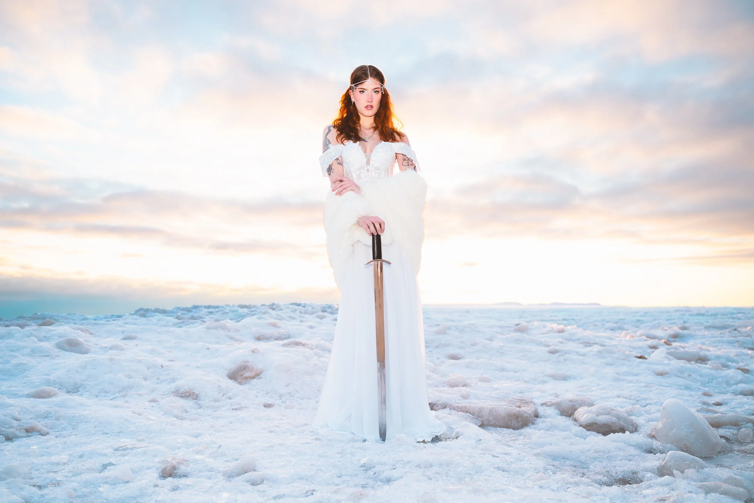 A woman dressed in a white gown holding a sword over snow, with a cloudy sky in the background.