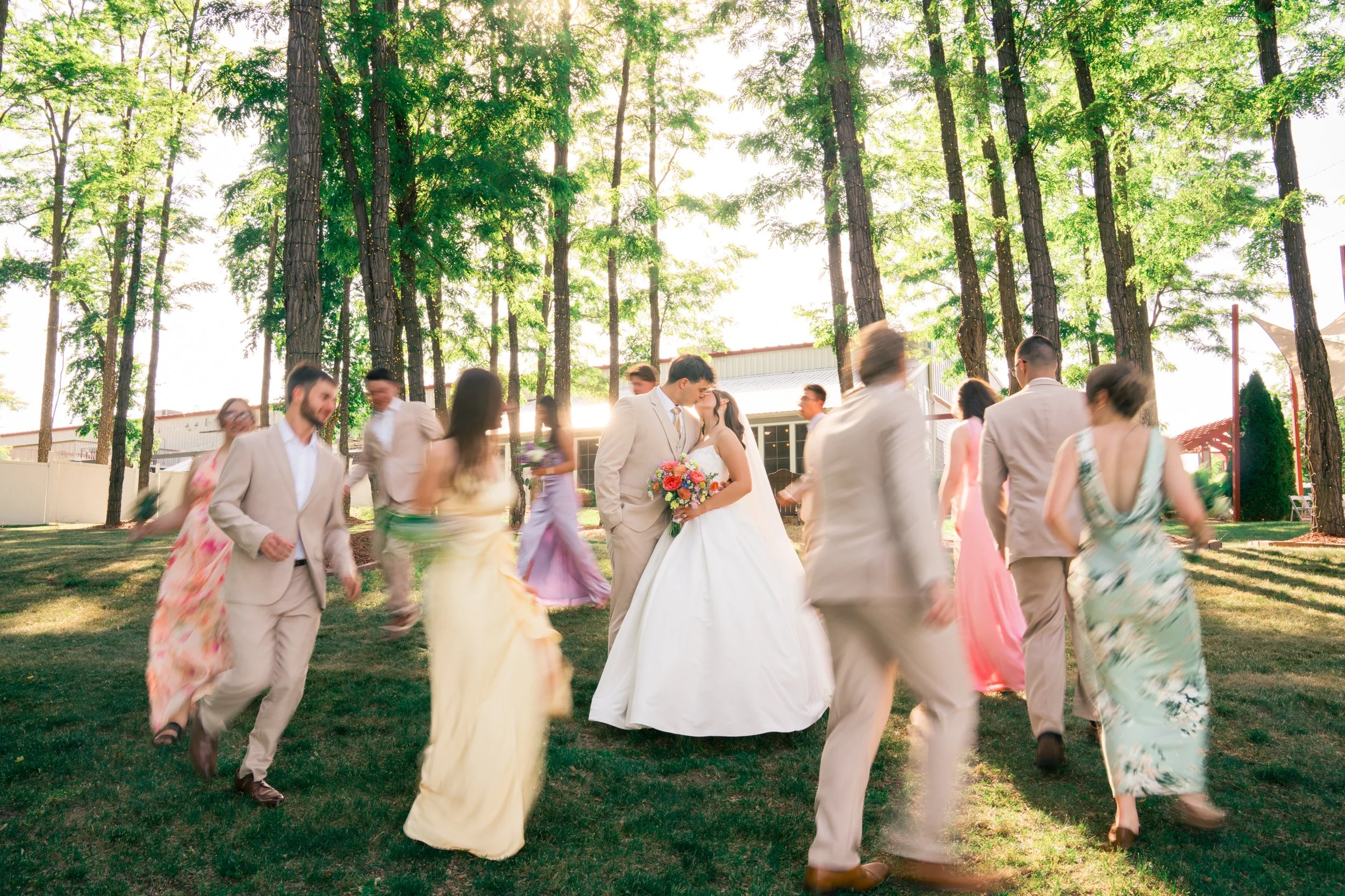 Bride and groom kissing in the center of a group of people dancing outdoors during a wedding celebration in a wooded area with sunlight filtering through the trees.