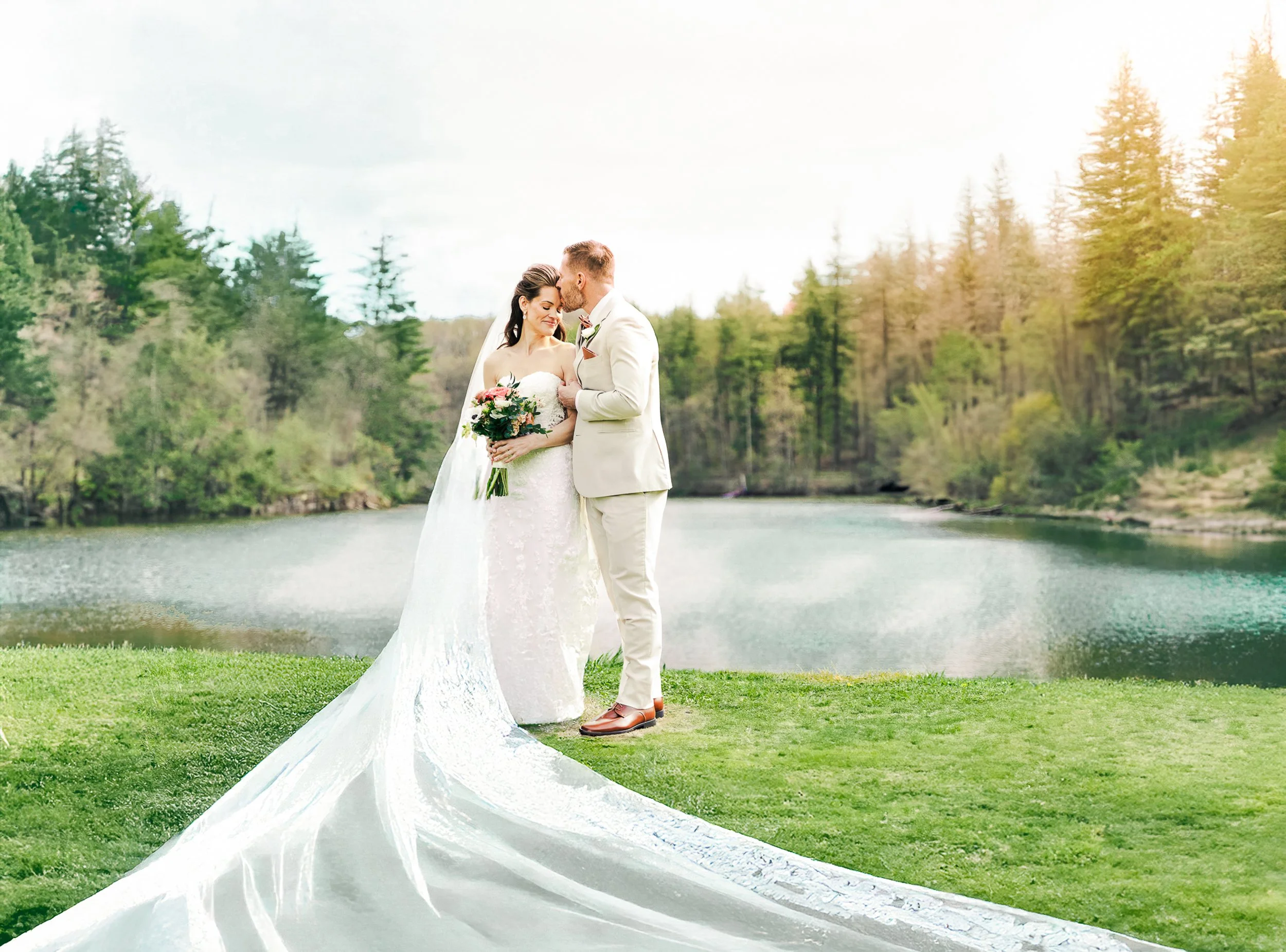 A bride and groom standing by a lake surrounded by trees, with the bride holding a bouquet and the groom leaning in for a kiss.