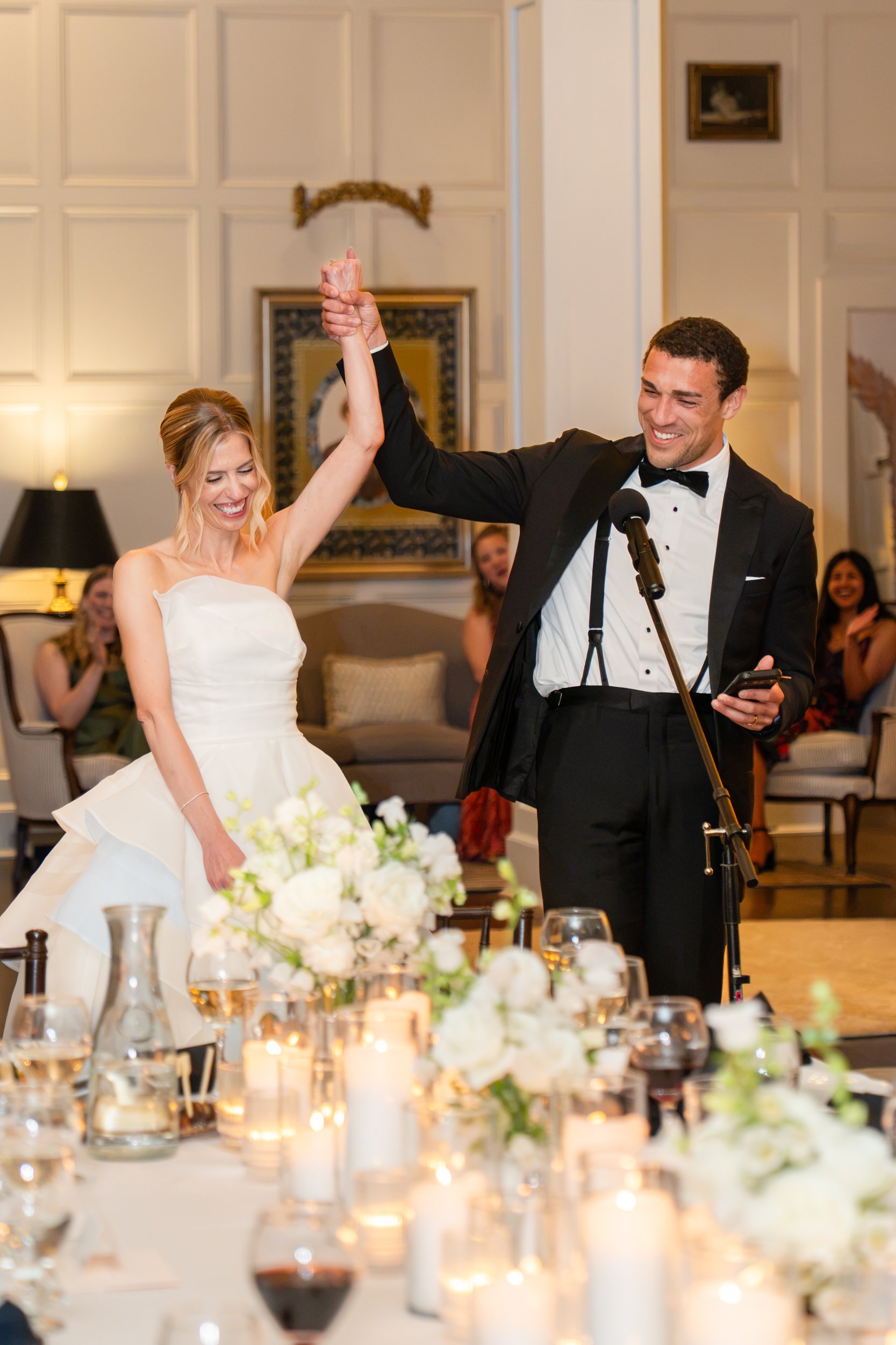 A newlywed bride and groom are celebrating at their wedding reception, holding hands in the air and smiling. The bride is wearing a strapless white wedding gown, and the groom is in a black tuxedo with a bow tie. Guests are seated in the background, 