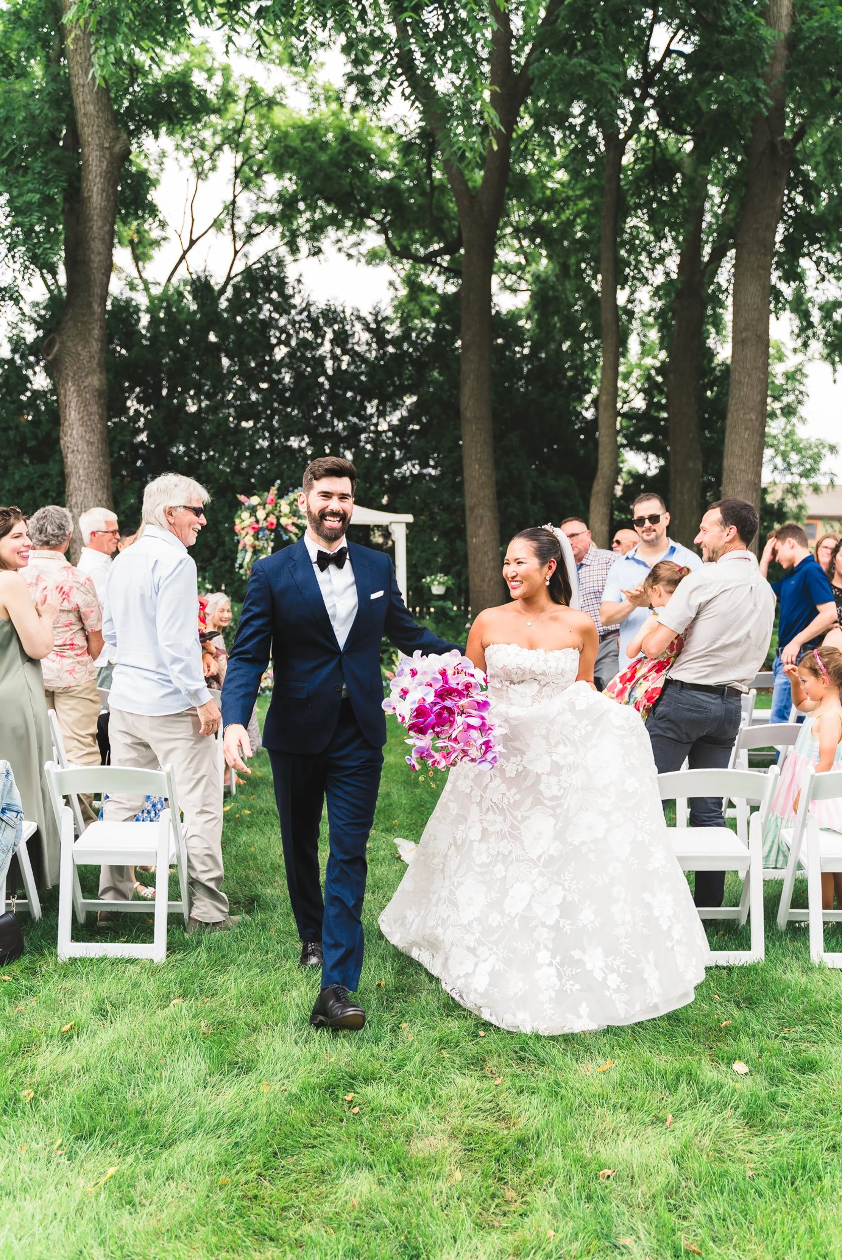 Happy bride and groom walking together outdoors at their wedding reception, surrounded by smiling guests, with green trees in the background.