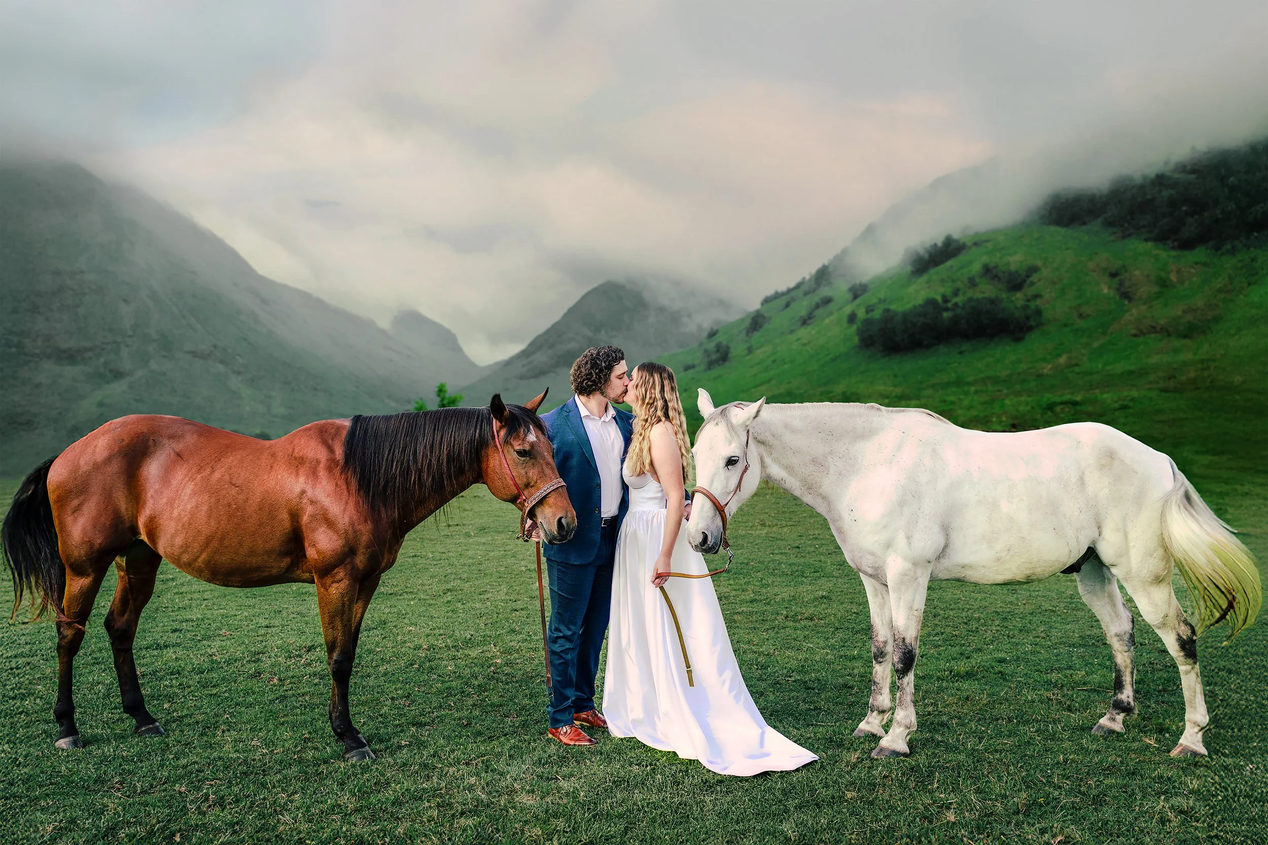 A couple in wedding attire kissing among three horses in a lush green mountain landscape, with misty mountains in the background.