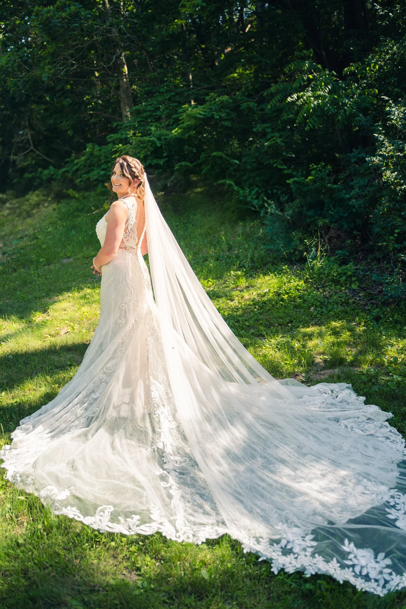 Bride in a white wedding dress with a long train, standing on grass in a lush green outdoor setting, smiling and looking over her shoulder.