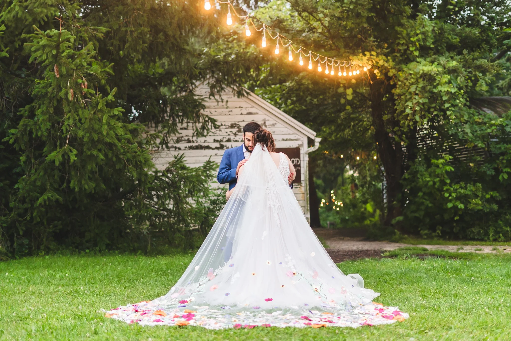 A bride and groom embrace outdoors during their wedding, with the bride wearing a long, white, floral-embellished veil and gown, and the groom in a blue suit. String lights hang above them, and there are trees and a small white building in the backgr