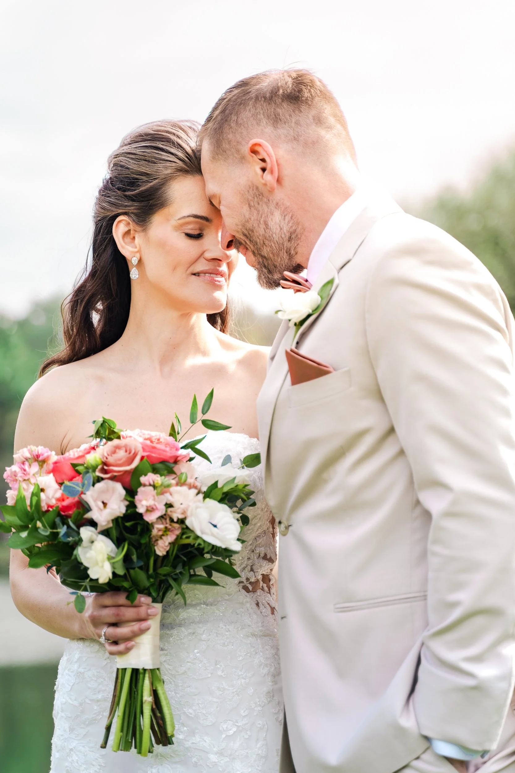 A bride and groom with foreheads touching, smiling with eyes closed, outdoors on their wedding day. The bride holds a bouquet of pink, white, and green flowers, wearing a strapless lace wedding dress. The groom wears a cream-colored suit, pink tie, a