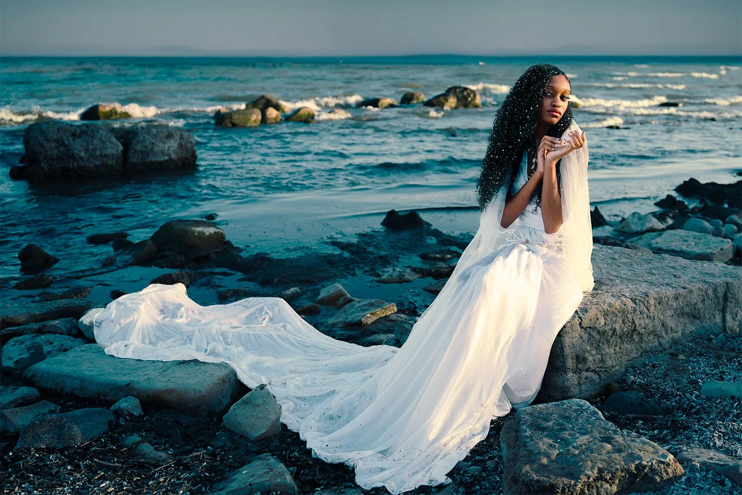 A woman in a white flowing dress sits on a large rock on a rocky beach by the ocean, holding her hands near her face, with long curly dark hair and a calm expression.