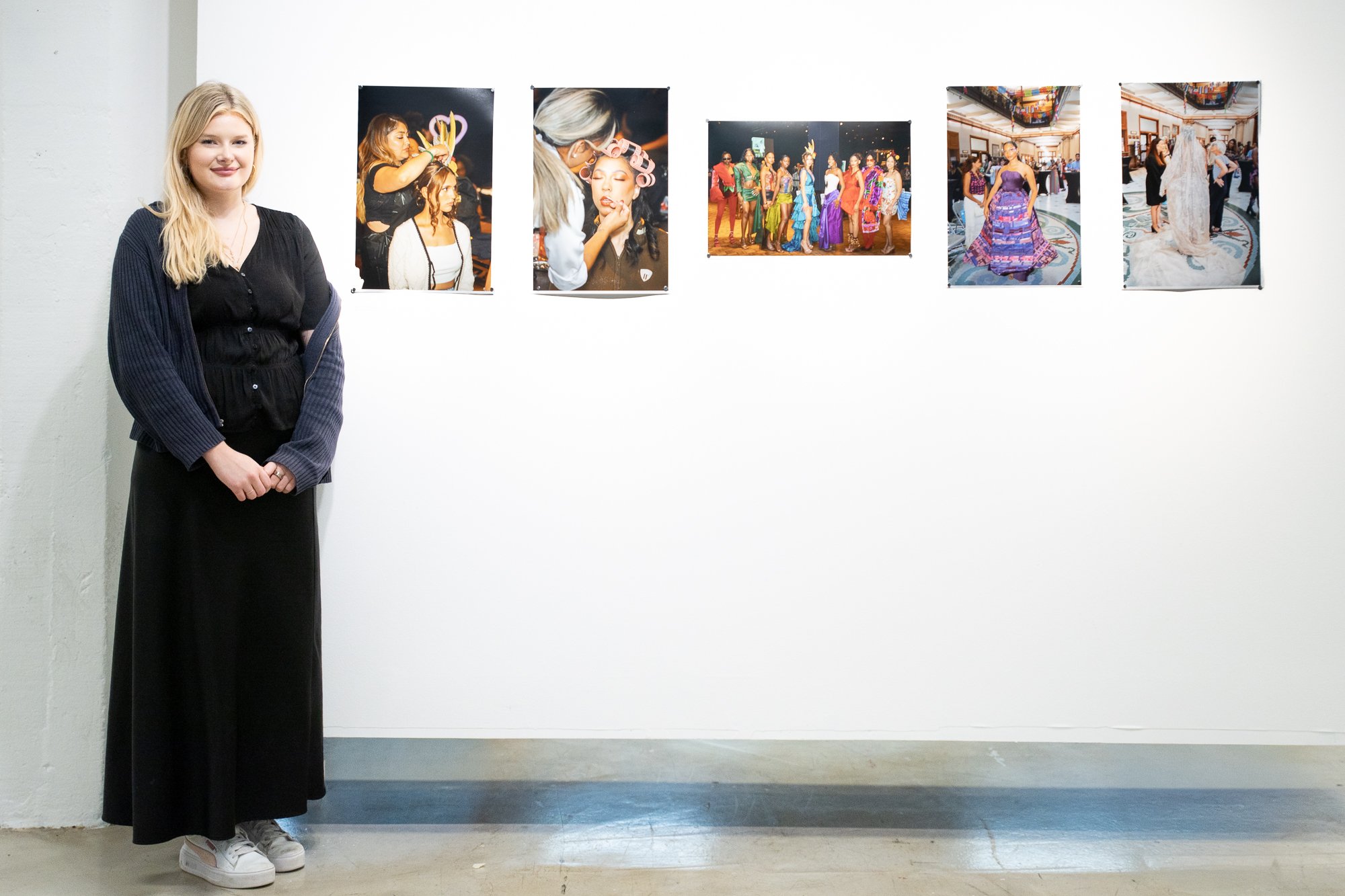 A woman with blonde hair wearing a black dress and a dark cardigan stands in an art gallery, next to a white wall displaying six large photos of a fashion show or event.