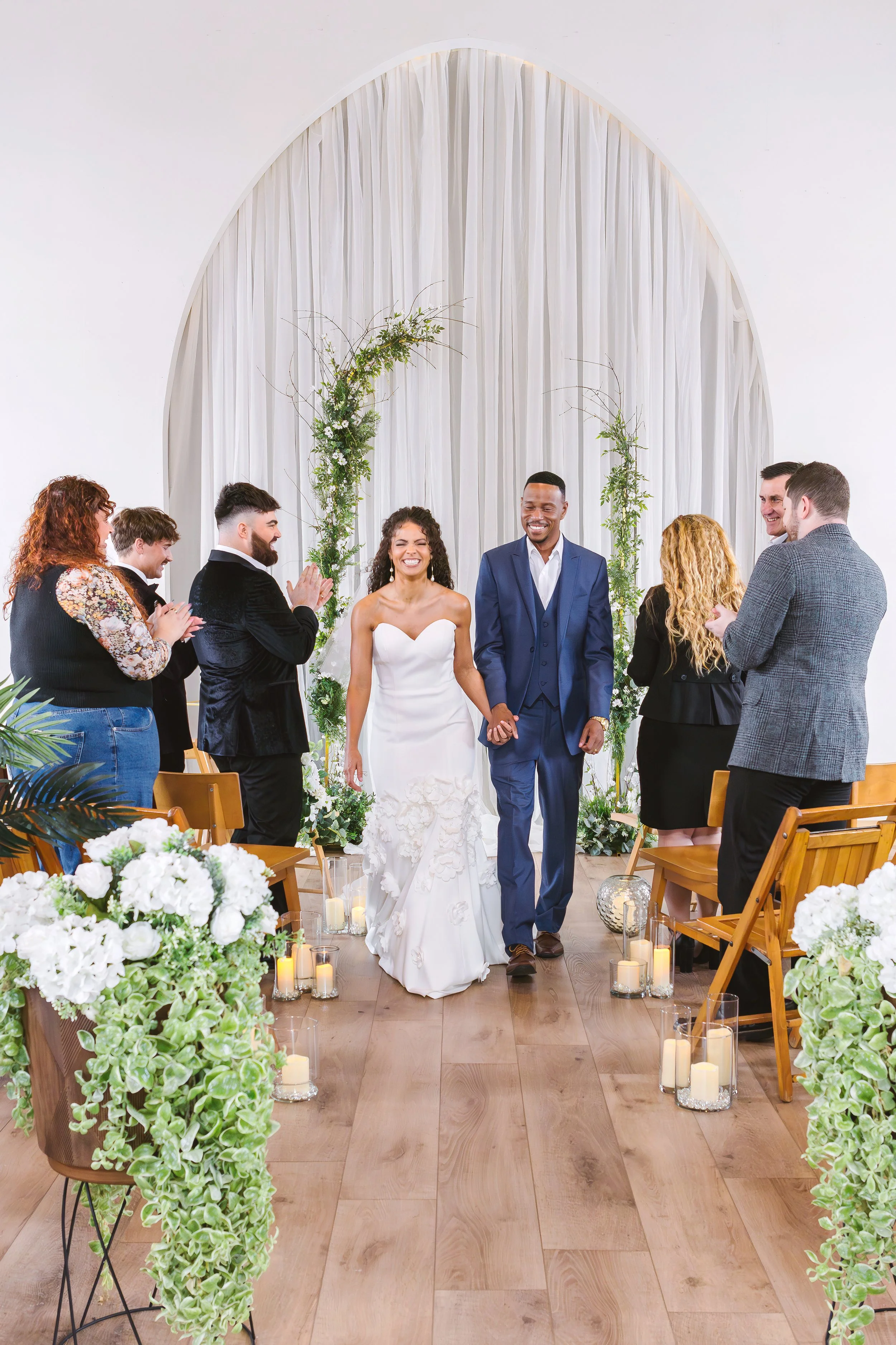 A wedding ceremony with a bride and groom walking hand in hand down the aisle, surrounded by friends and family, inside a decorated venue with white drapes, green floral arrangements, and candles.