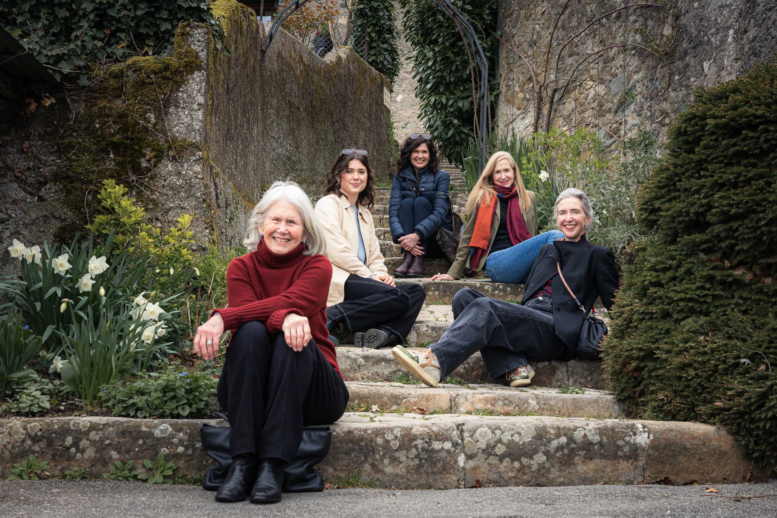 Cinq femmes assises sur des escaliers en pierre dans un jardin avec des fleurs et des plantes.