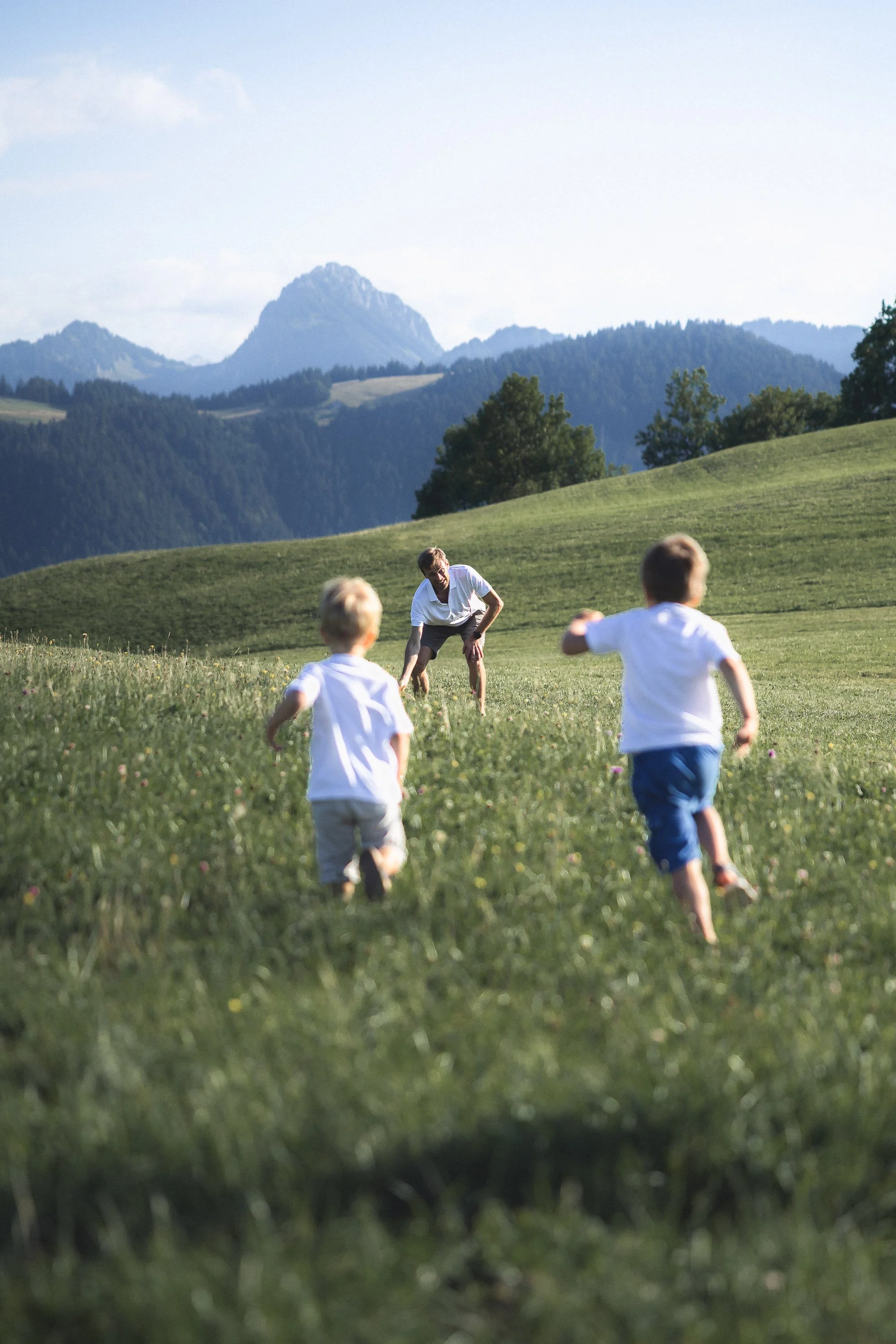 Photo de famille en Haute-Savoie par le photographe Rüdi Welter. Deux enfants qui cours vers leur père dans des herbes haute avec une montagne en arrière-plan.