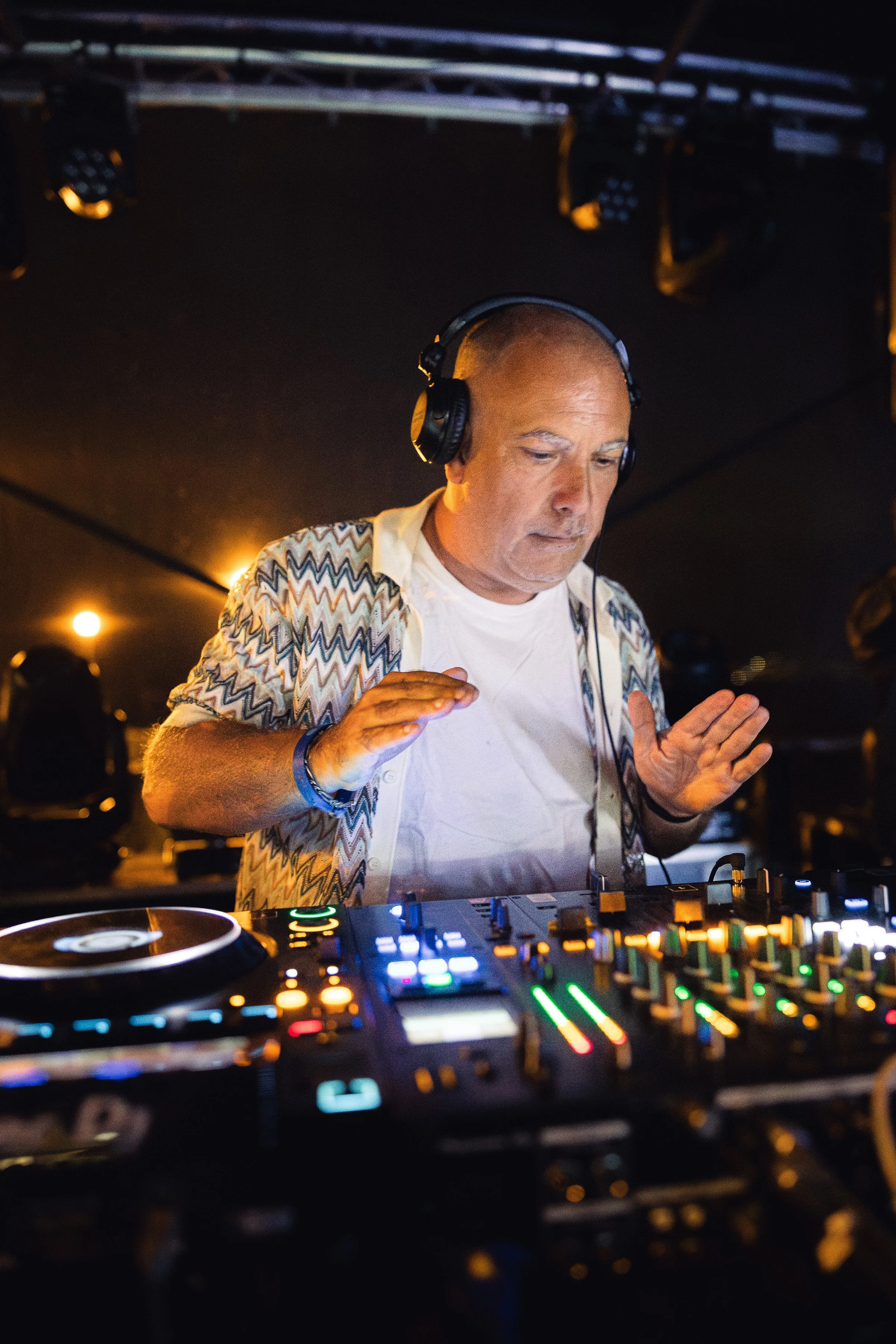 Un DJ portant un casque noir, avec une chemise à motifs zigzag, joue de la musique dans un club ou une fête, avec des contrôleurs DJ lumineux devant lui, en ambiance sombre et illuminée par des lumières chaudes.