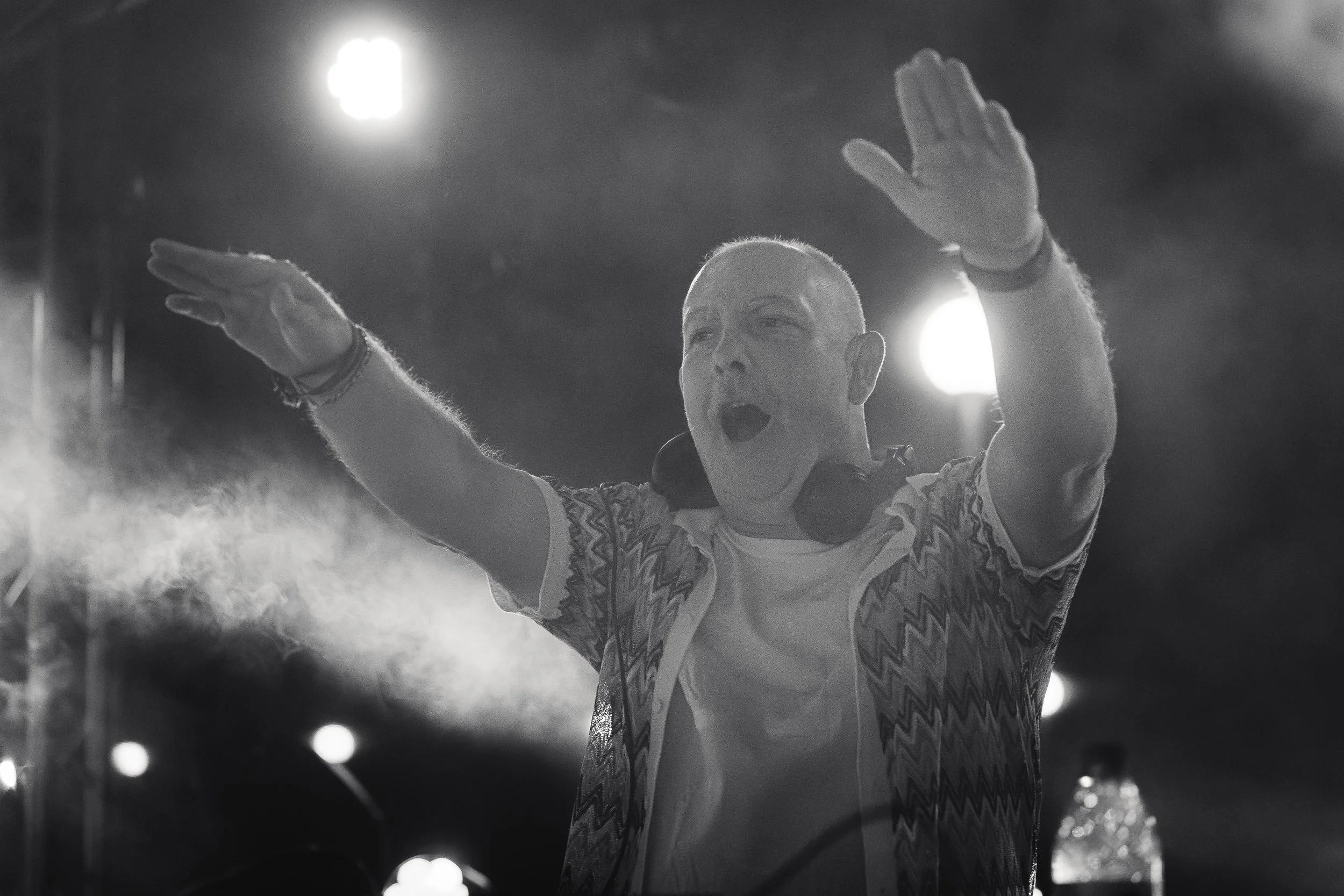 Un DJ avec des écouteurs autour du cou, les bras levés, semble en train de performer devant un public, éclairage de scène en fond.