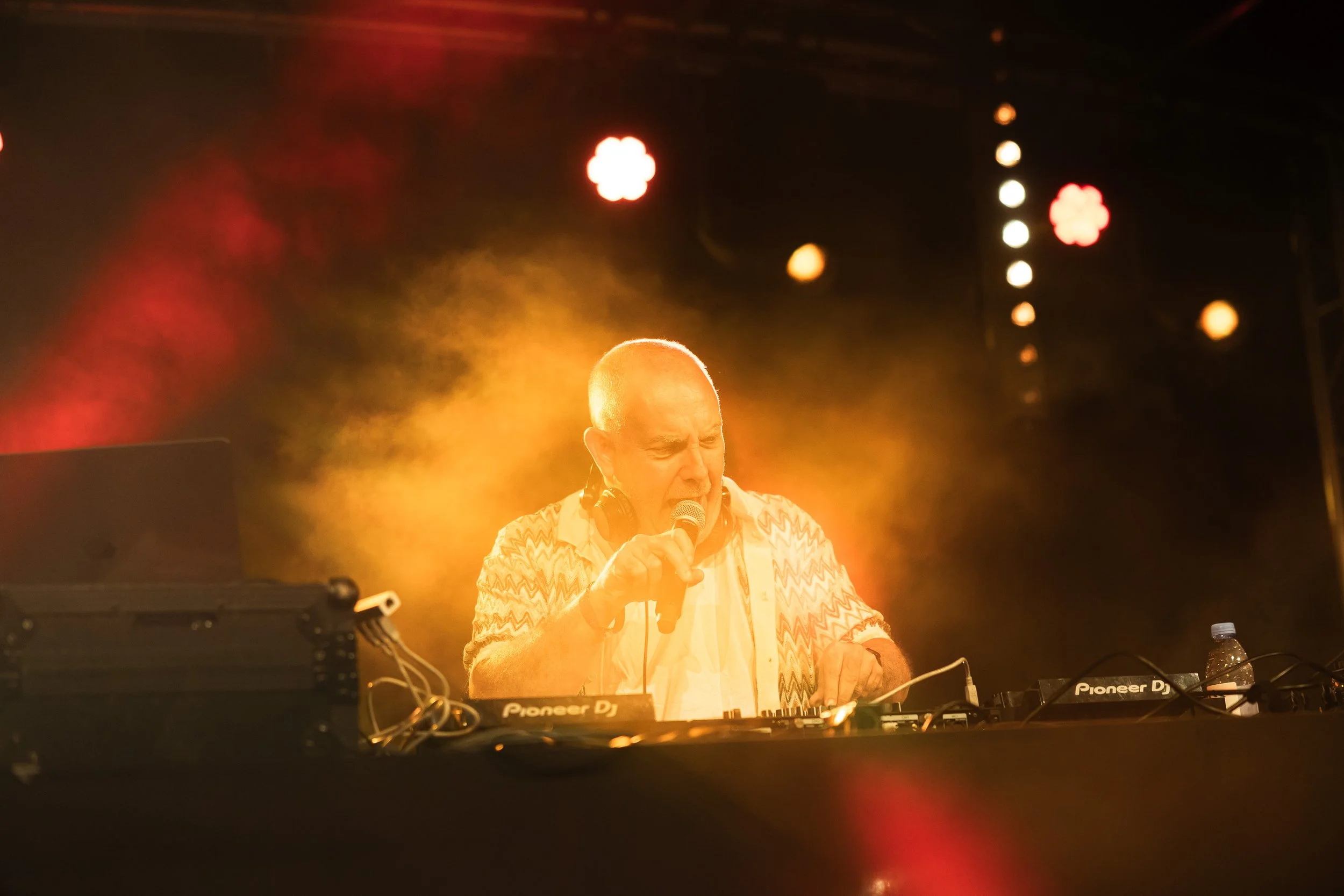 DJ homme avec une veste à motif, chantant dans un microphone lors d'une performance sur scène, entouré d'équipements de musique et sous un éclairage chaud orange.