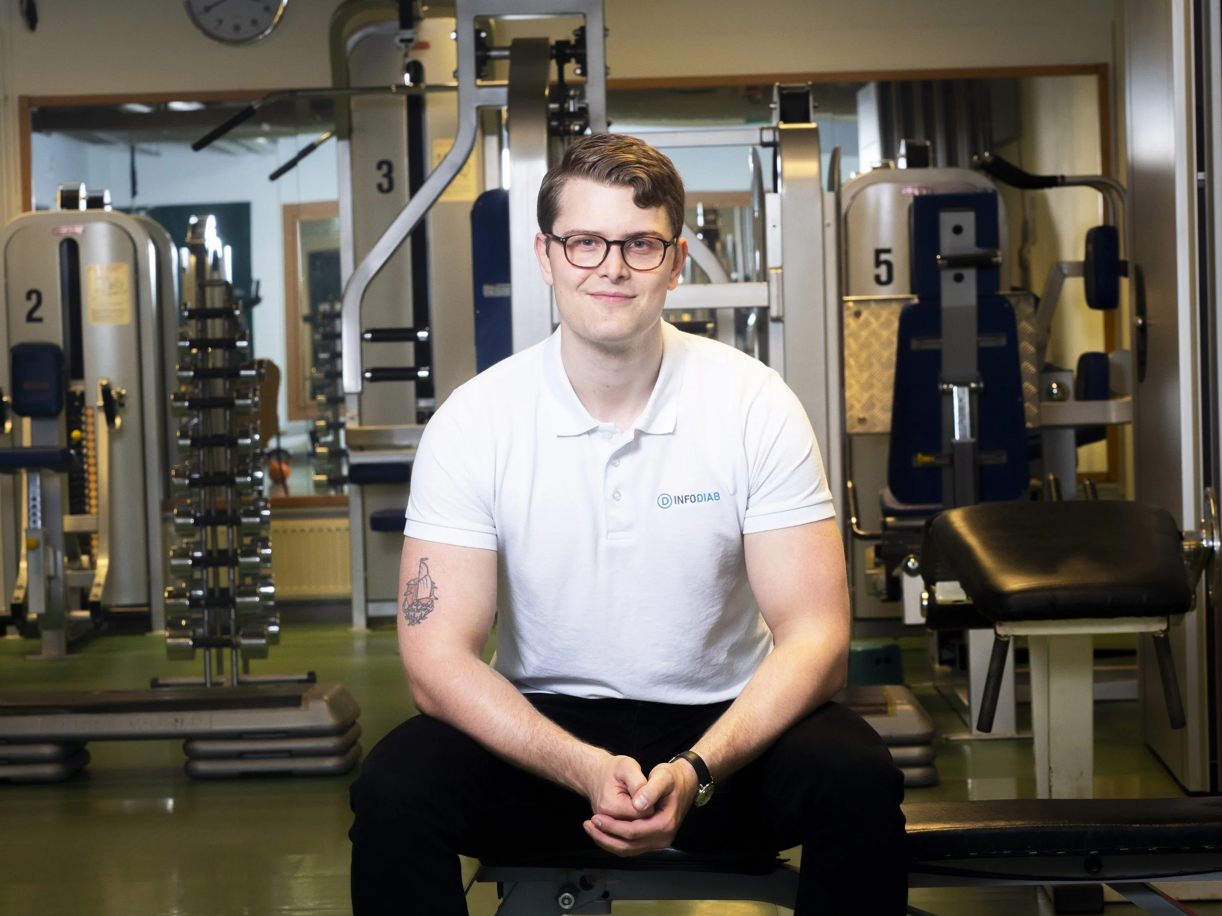 En man i vit piké sitter på en träningsbänk i ett välutrustat gym.