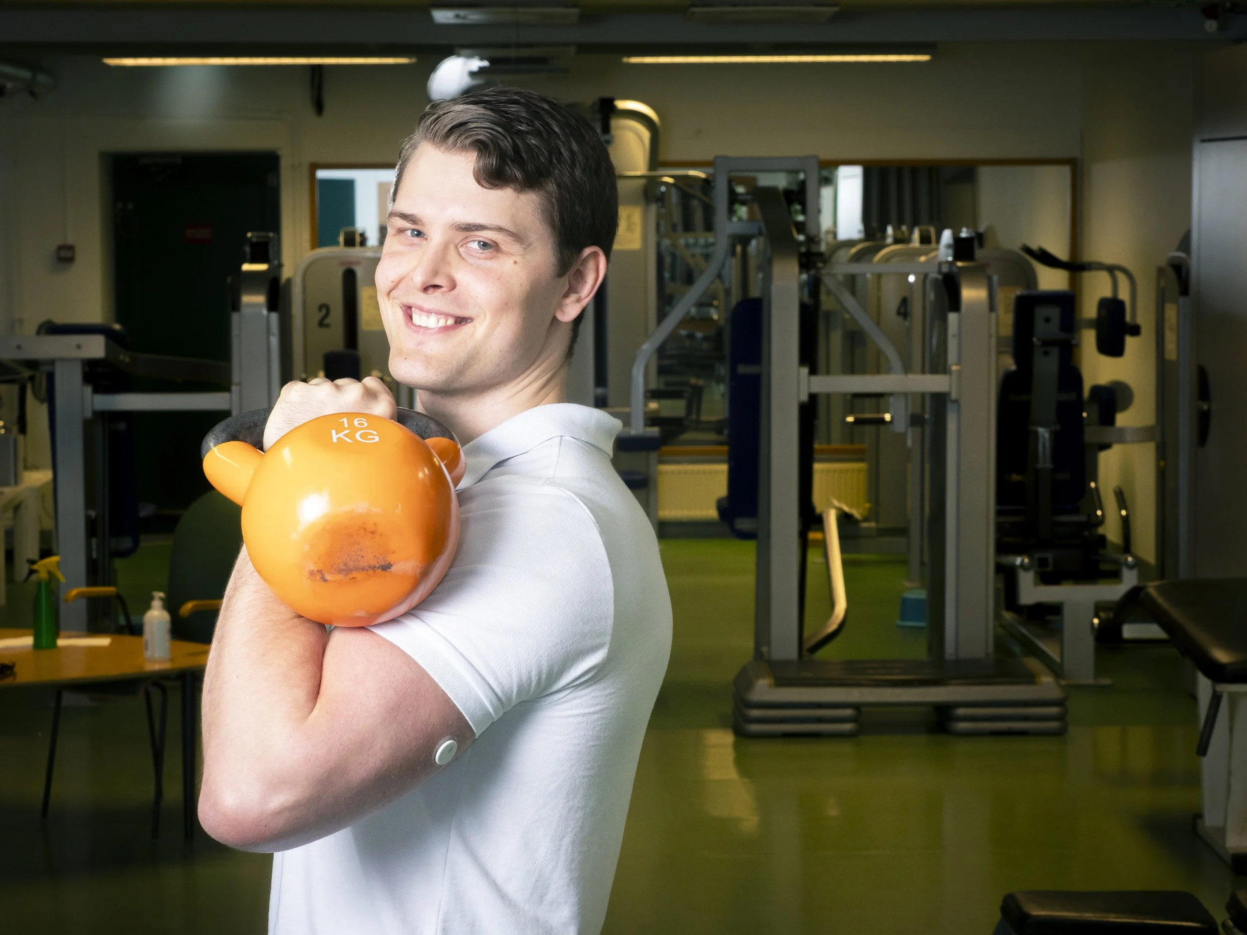 En ung man som håller en orange kettlebell med vikten 16 kg framför en gymmiljö.