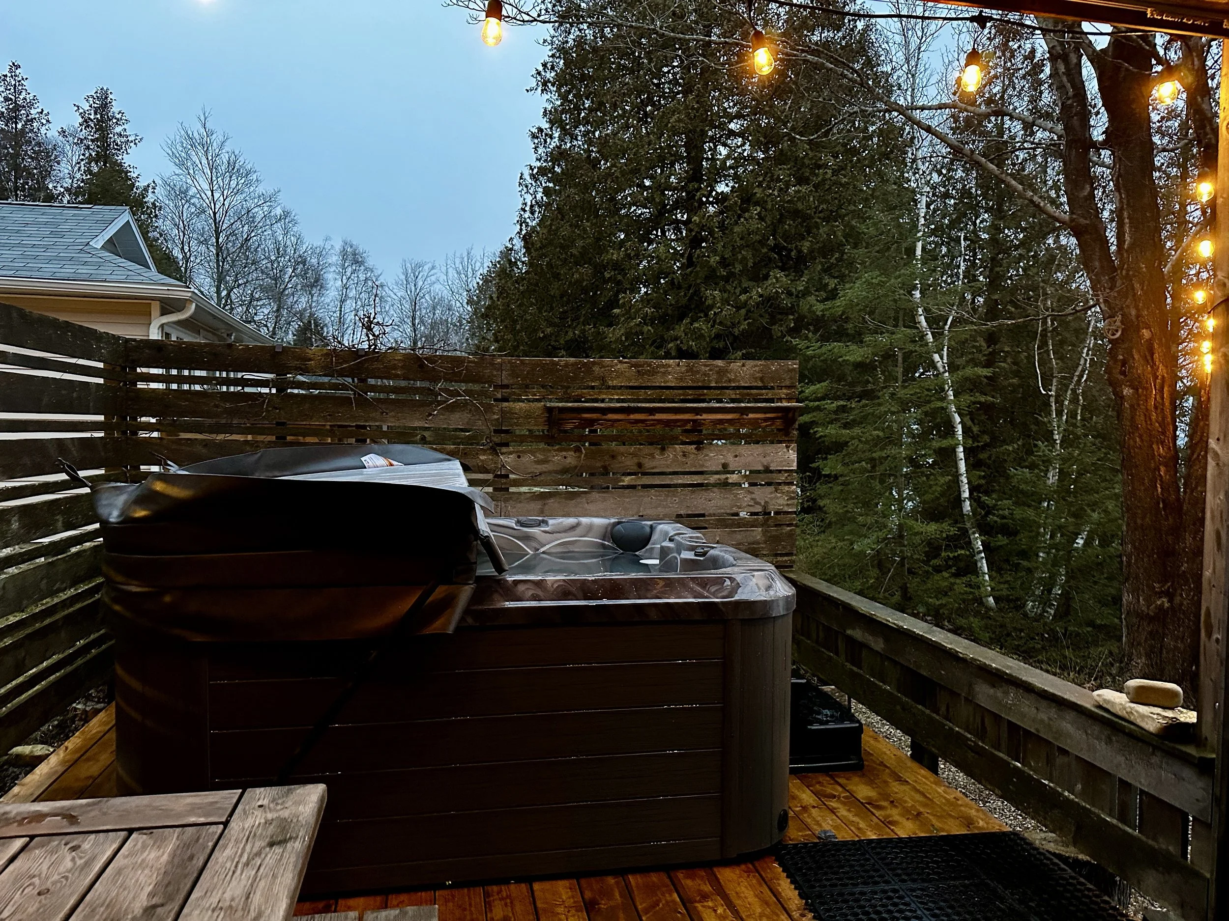 An outdoor hot tub on a wooden deck, surrounded by a wooden fence and string lights, overlooking a wooded area during dusk.