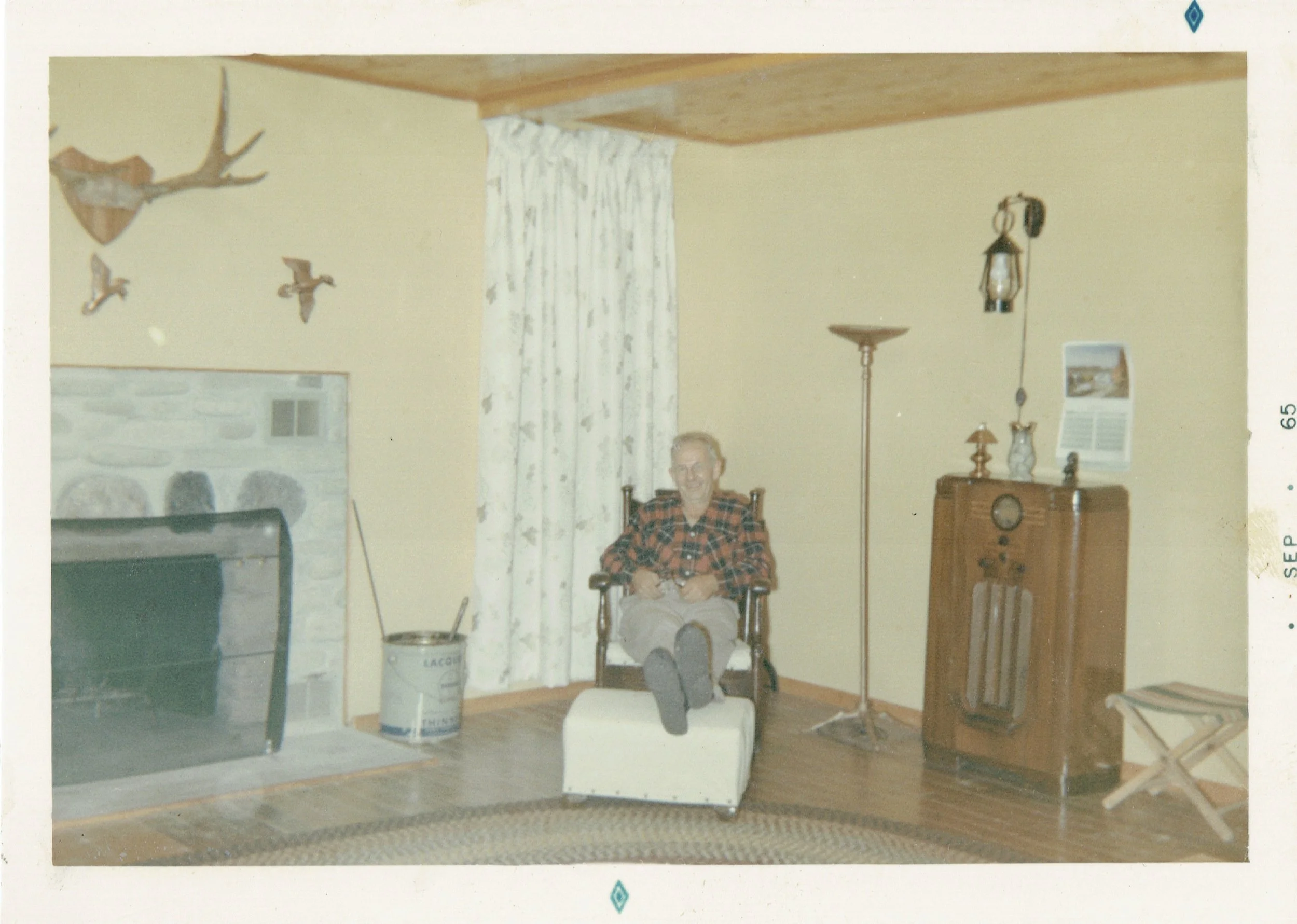 An elderly man sitting in a rocking chair in a living room with beige walls, a stone fireplace, a mounted deer head, wall-mounted bird decorations, a floor lamp, a vintage radio cabinet, and a floor-standing lamp.