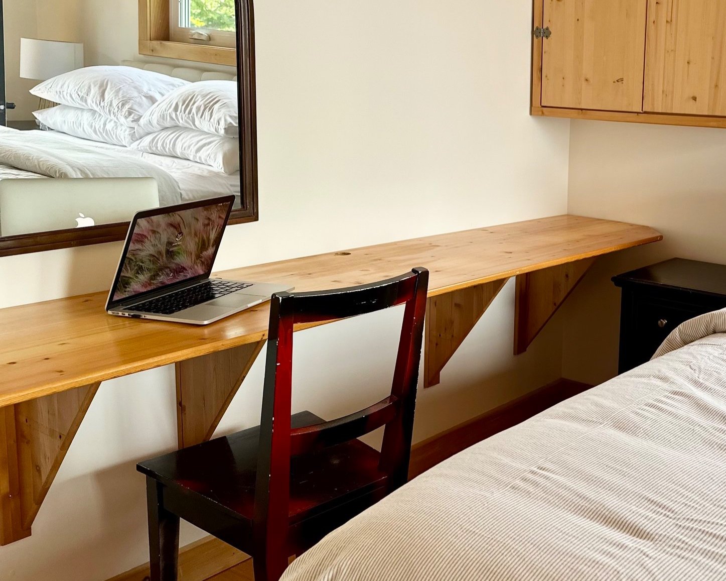 A wooden desk with a laptop, red and black chair, beige bed with striped blanket, and a mirror reflecting a bedroom with a bed and window.