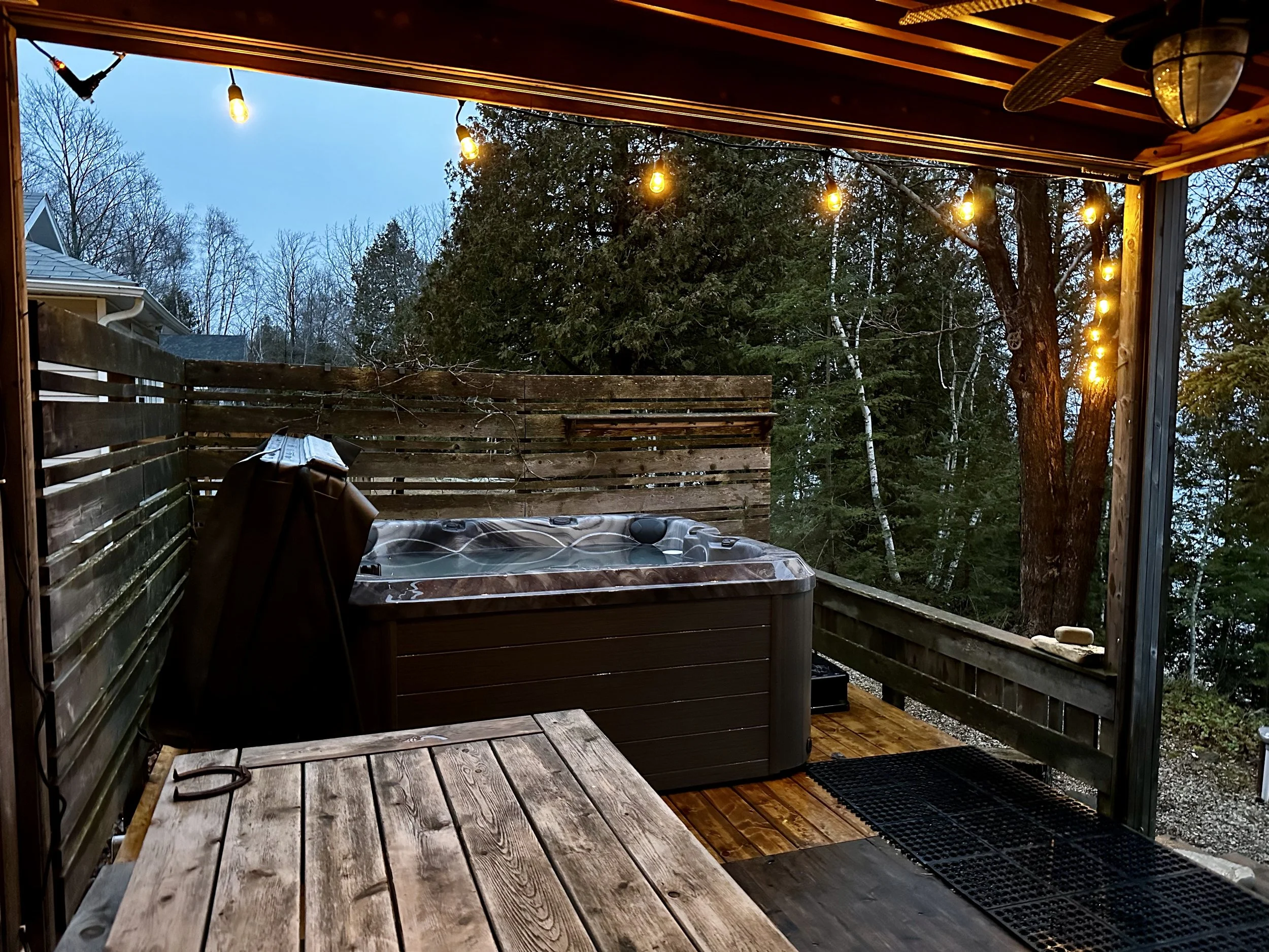 A cozy outdoor patio with a hot tub, string lights, a wooden table, and surrounded by trees.