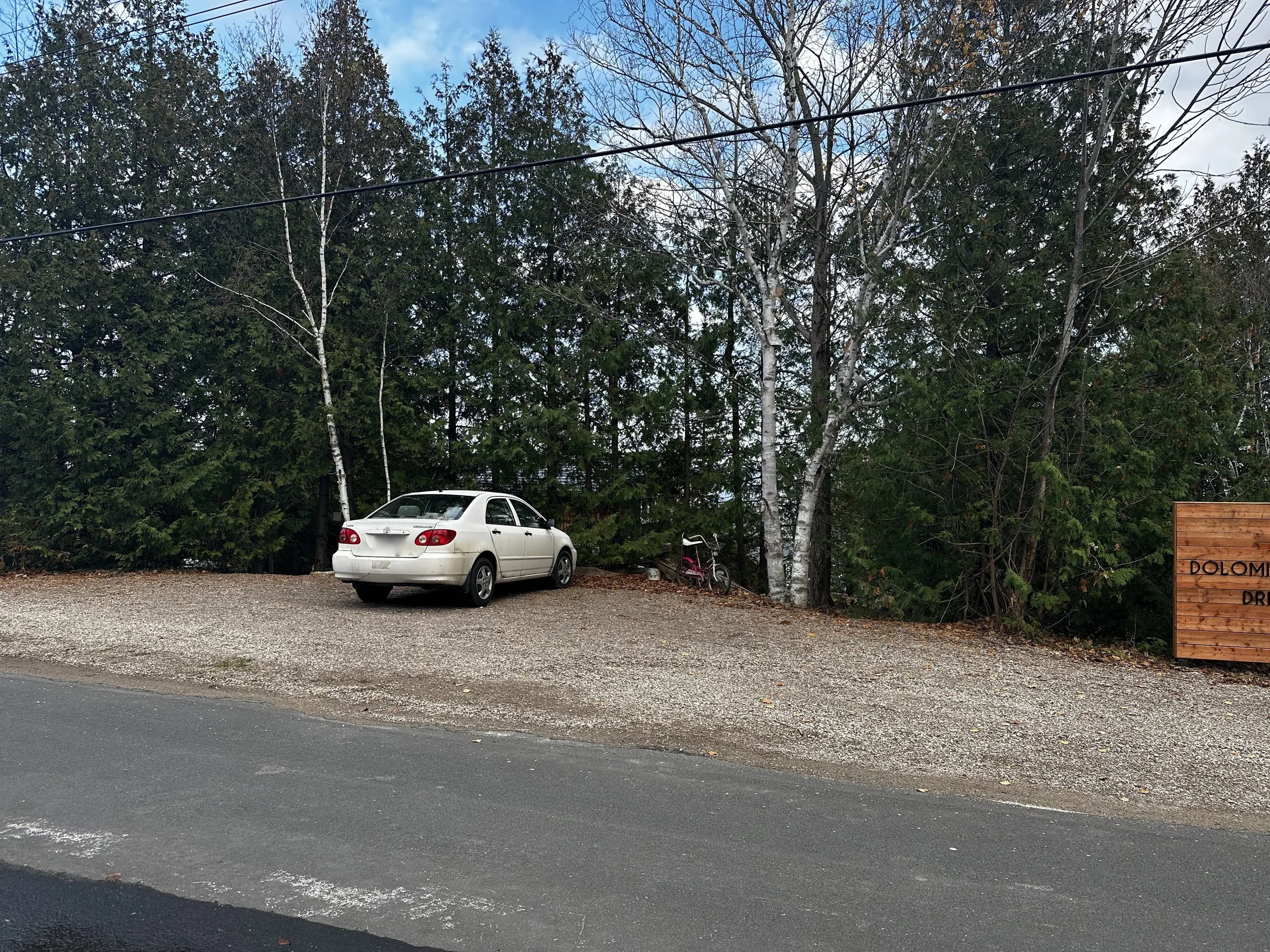 A white sedan parked on a gravel area beside a paved road, with a background of dense green trees and a partly cloudy sky.