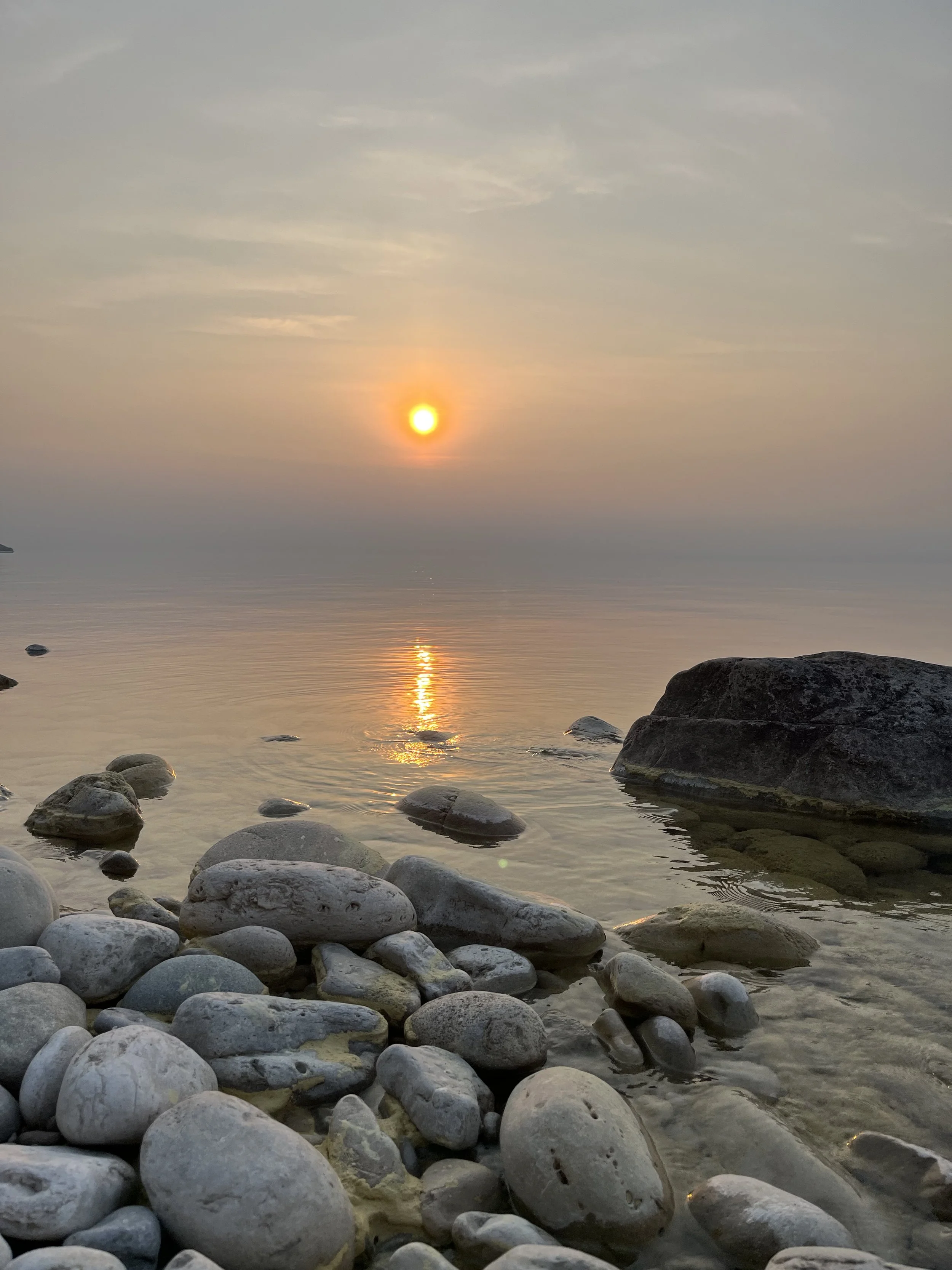 Sunset over calm water with rocks in the foreground and a sky with scattered clouds.