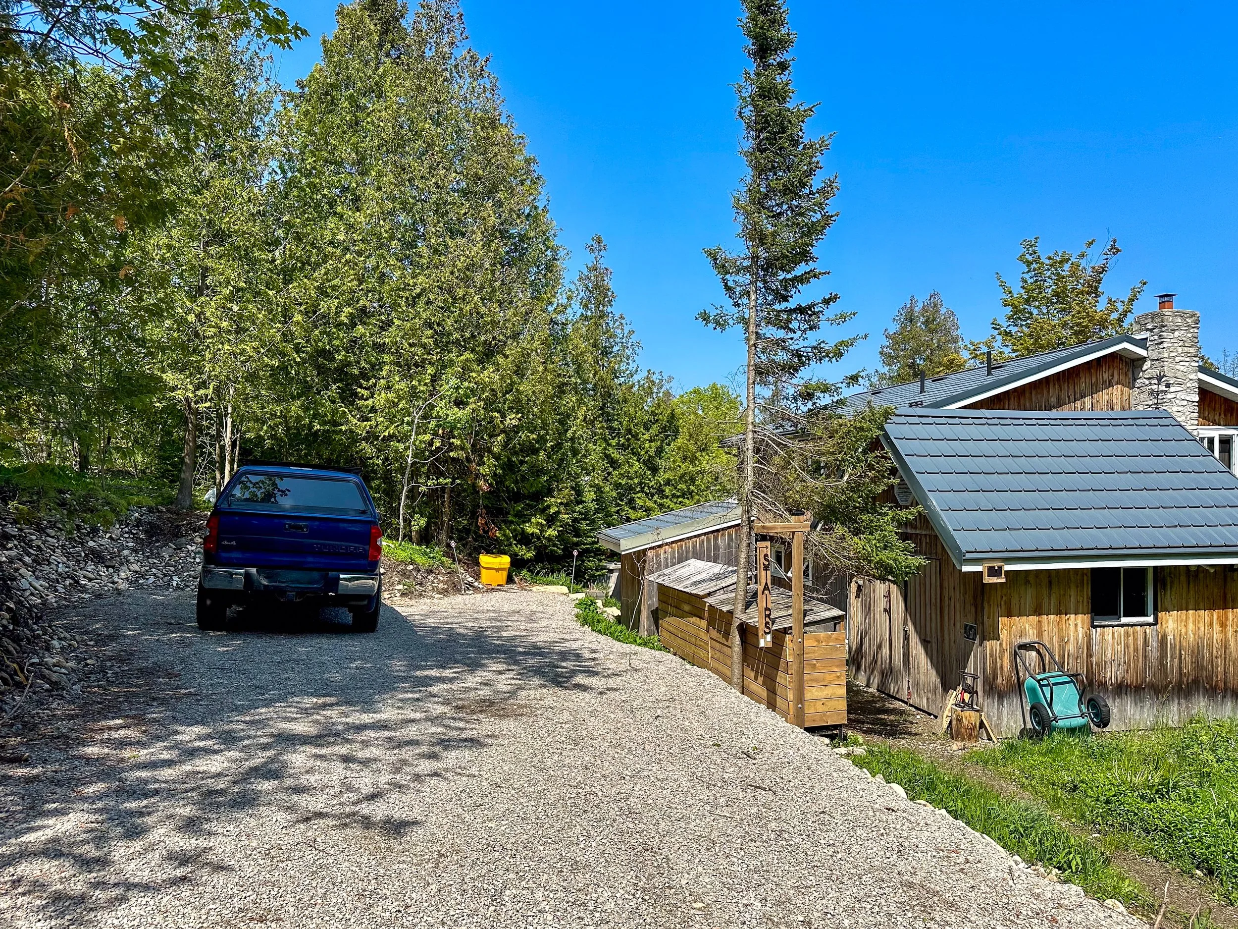 A gravel driveway leading to a wooden house with a metal roof, surrounded by trees. A blue pickup truck is parked on the driveway. There is a yellow container and a green wheelbarrow near the house.