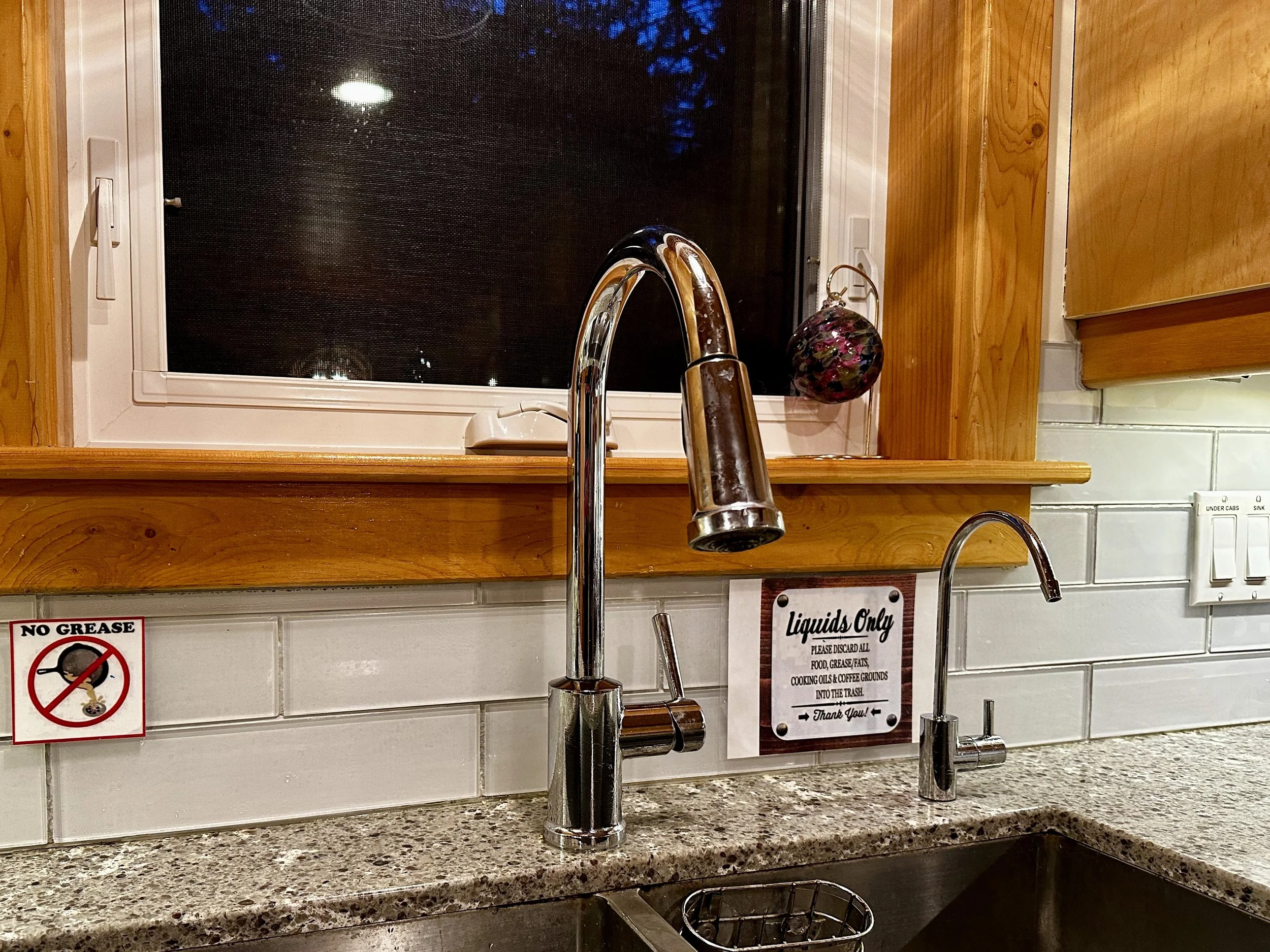 Kitchen counter with two sinks, one with a modern chrome faucet, and a window above. There are signs on the backsplash, one says "No Grease" with a crossed-out image of a grease glass, and the other says "Liquids Only" with instructions to discard all food, grease, fats, and cooking oils into the trash. A small pot or decorative object hangs by the window.