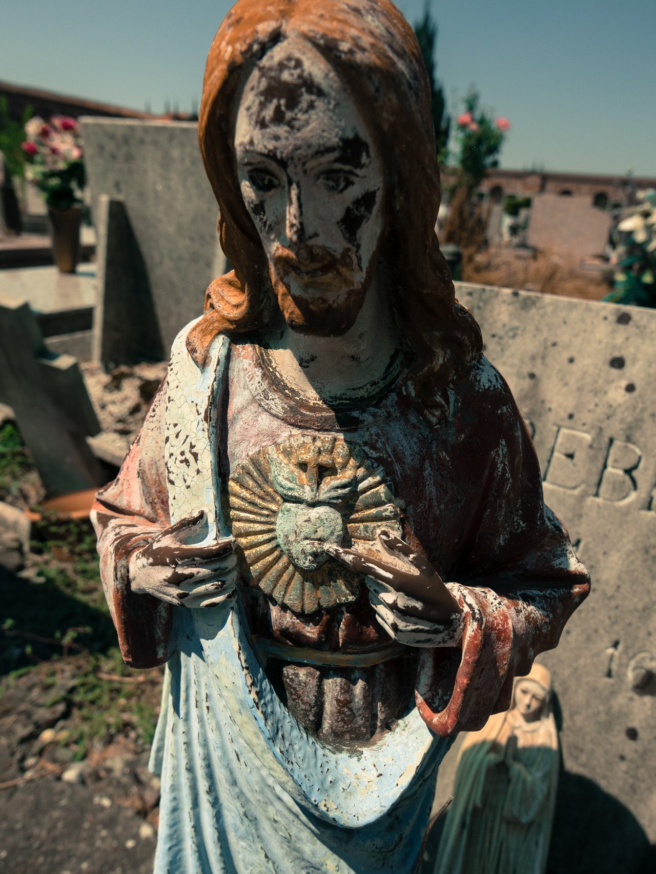 Weathered Jesus statue holding a heart with a cross inside a sunburst design, in a cemetery.