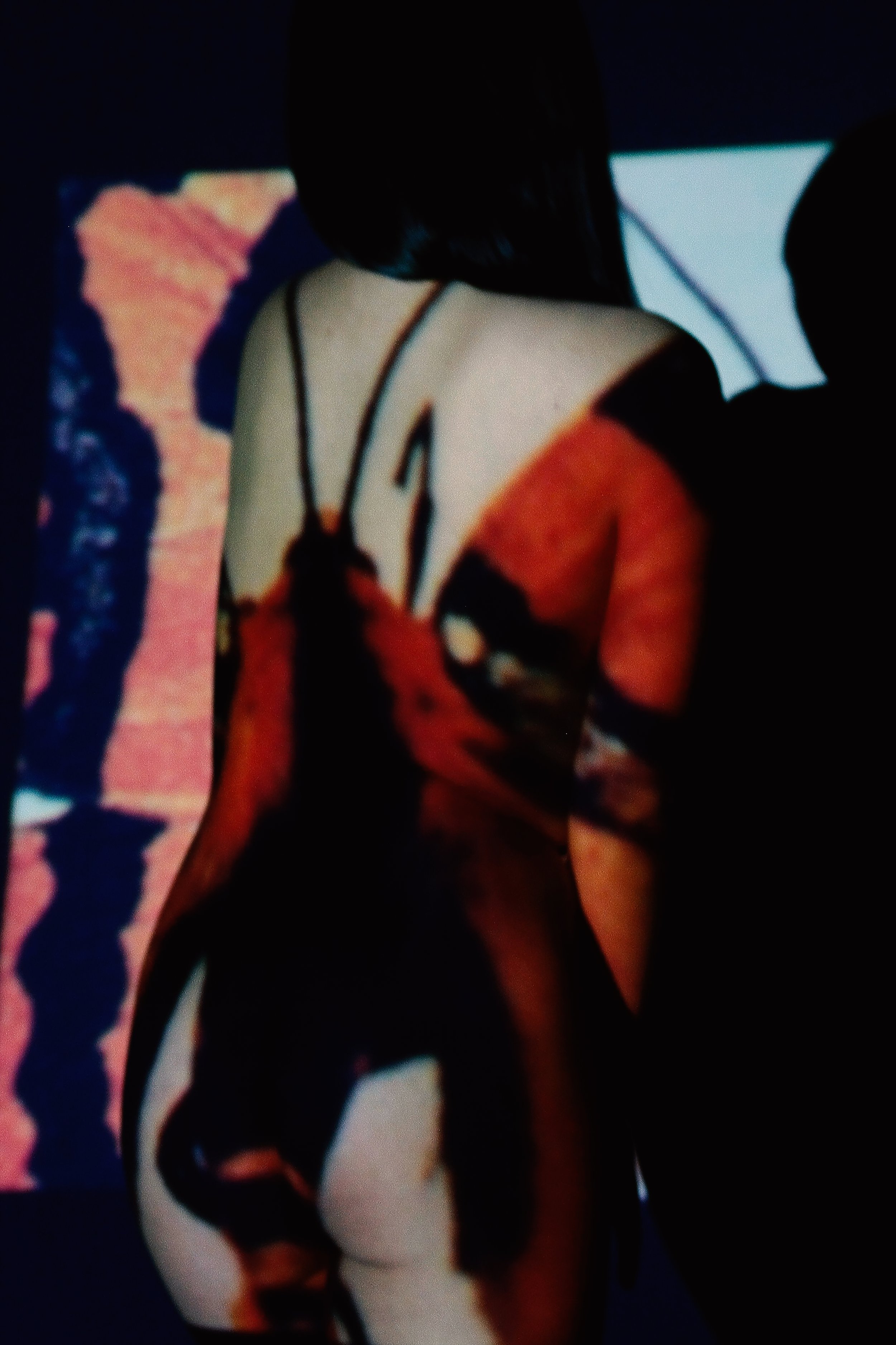 Person with long dark hair wearing a spaghetti strap dress with a dark and orange pattern, standing in front of a screen displaying abstract images.