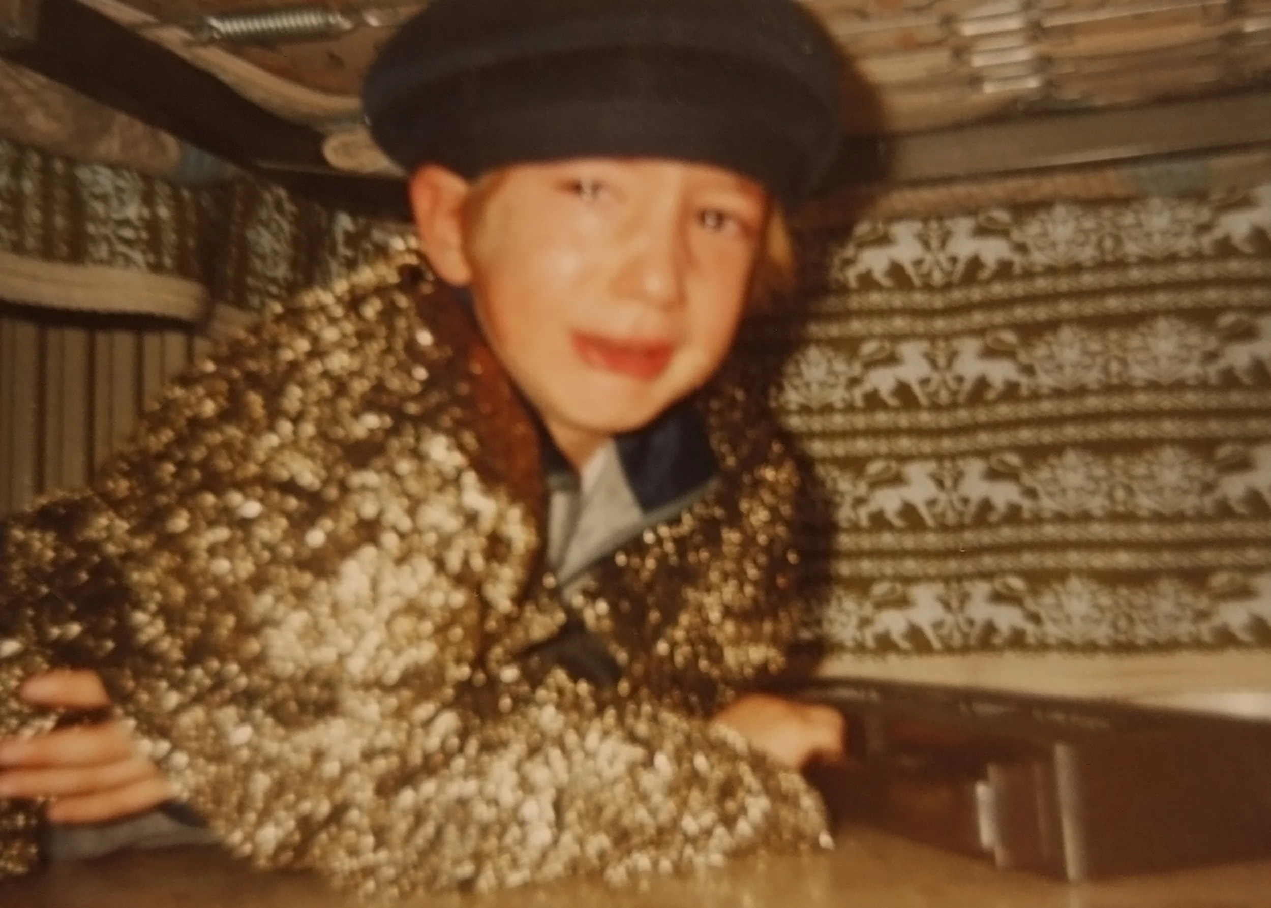 A person wearing a black hat and a gold, textured jacket, leaning on a wooden surface, with a decorative patterned wall in the background.