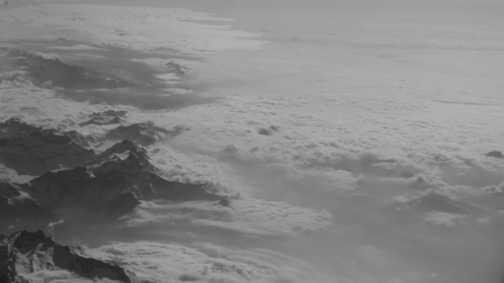Aerial view of snow-capped mountain peaks above clouds, looking down from high altitude.