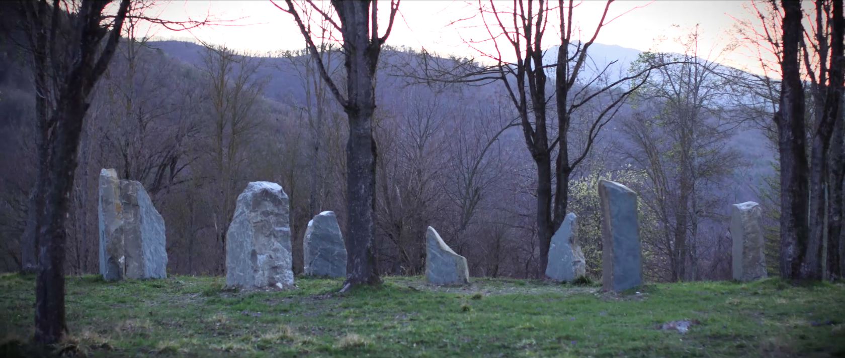 A circle of tall, irregular stone standing stones set in a grassy clearing amidst leafless trees with mountains in the background during dusk.
