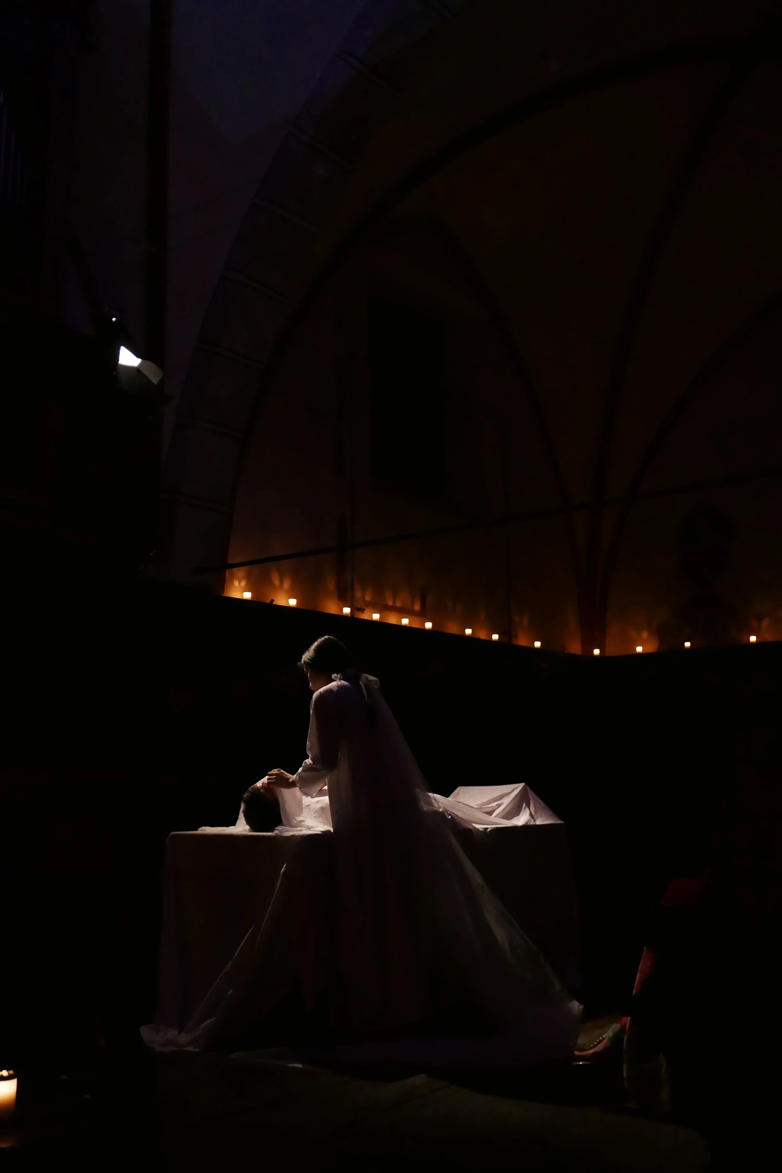 A woman in a white dress preparing a body for a funeral or burial in a dimly lit church or chapel with candles.