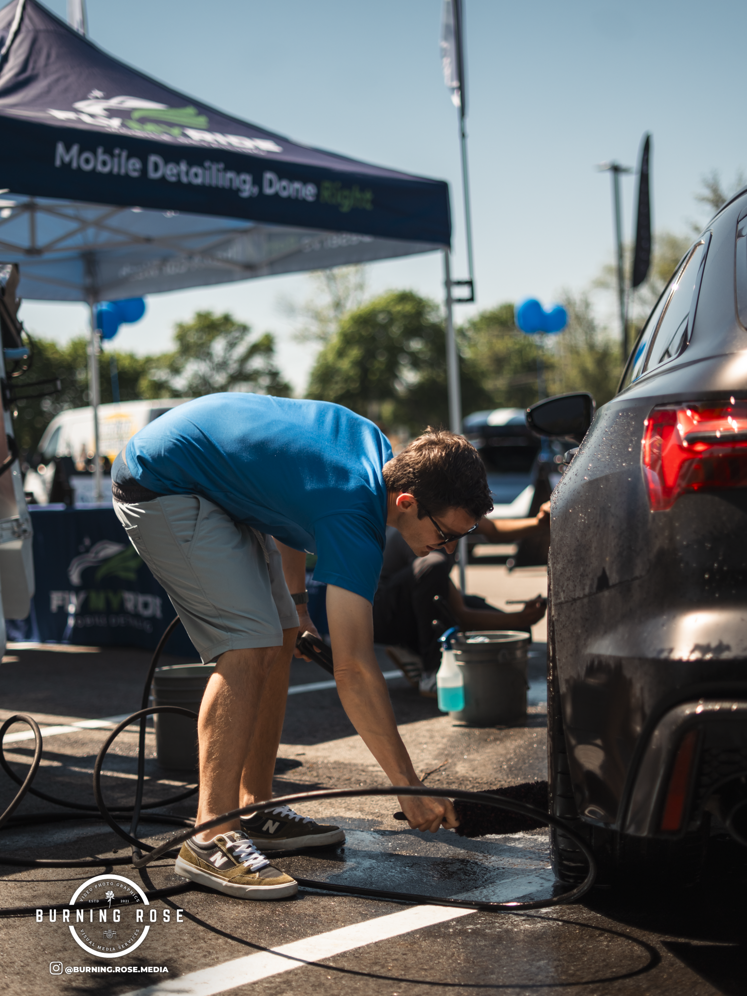A man in a blue shirt and shorts is cleaning the back of a black car at an outdoor event with a blue canopy in the background. The scene is sunny with trees in the distance.