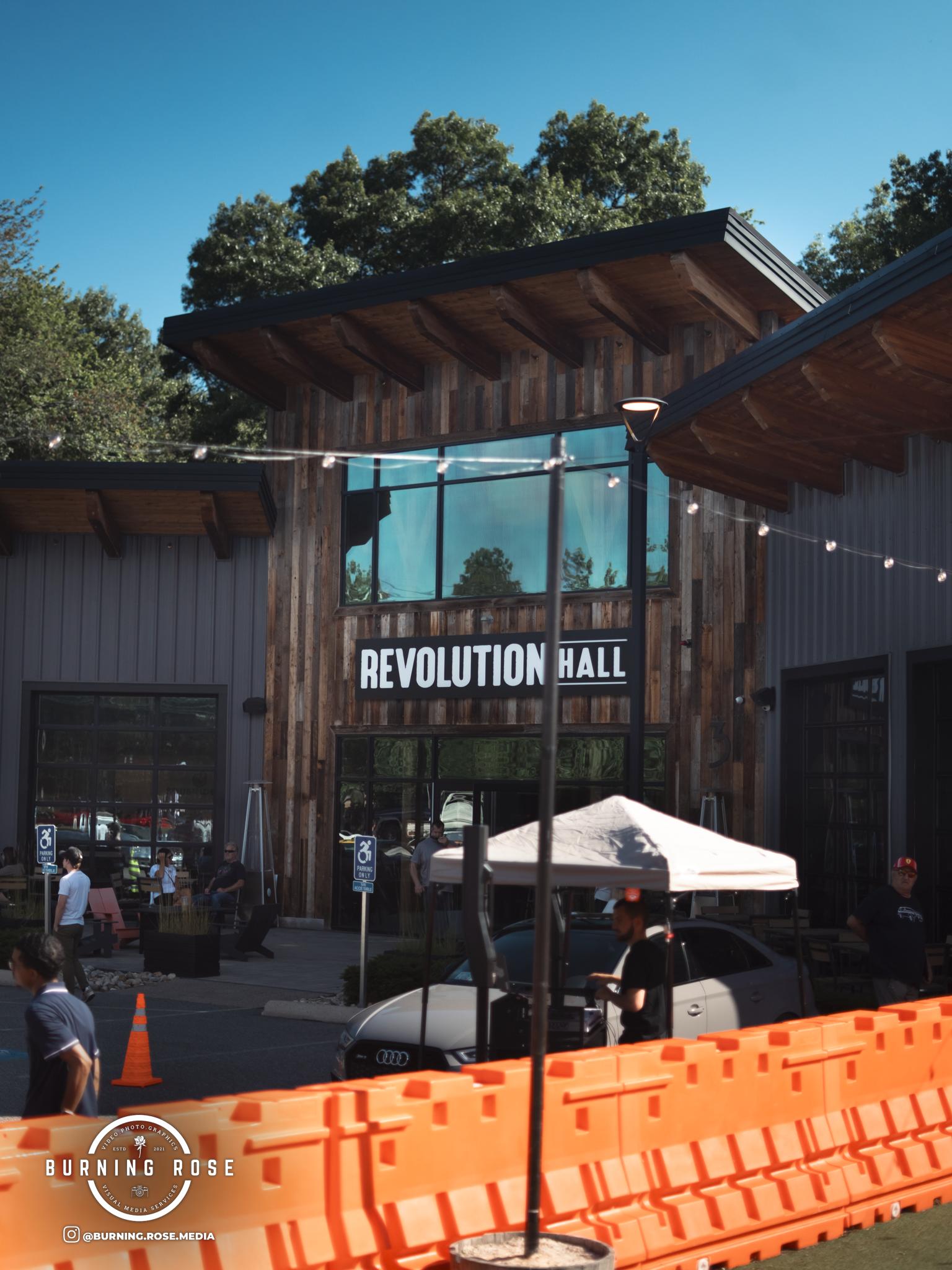 People gathered outside a wooden building with a sign that reads 'Revolution Hall.' Orange barricades and caution cones are visible, and some individuals are sitting or standing near a tent and cars.