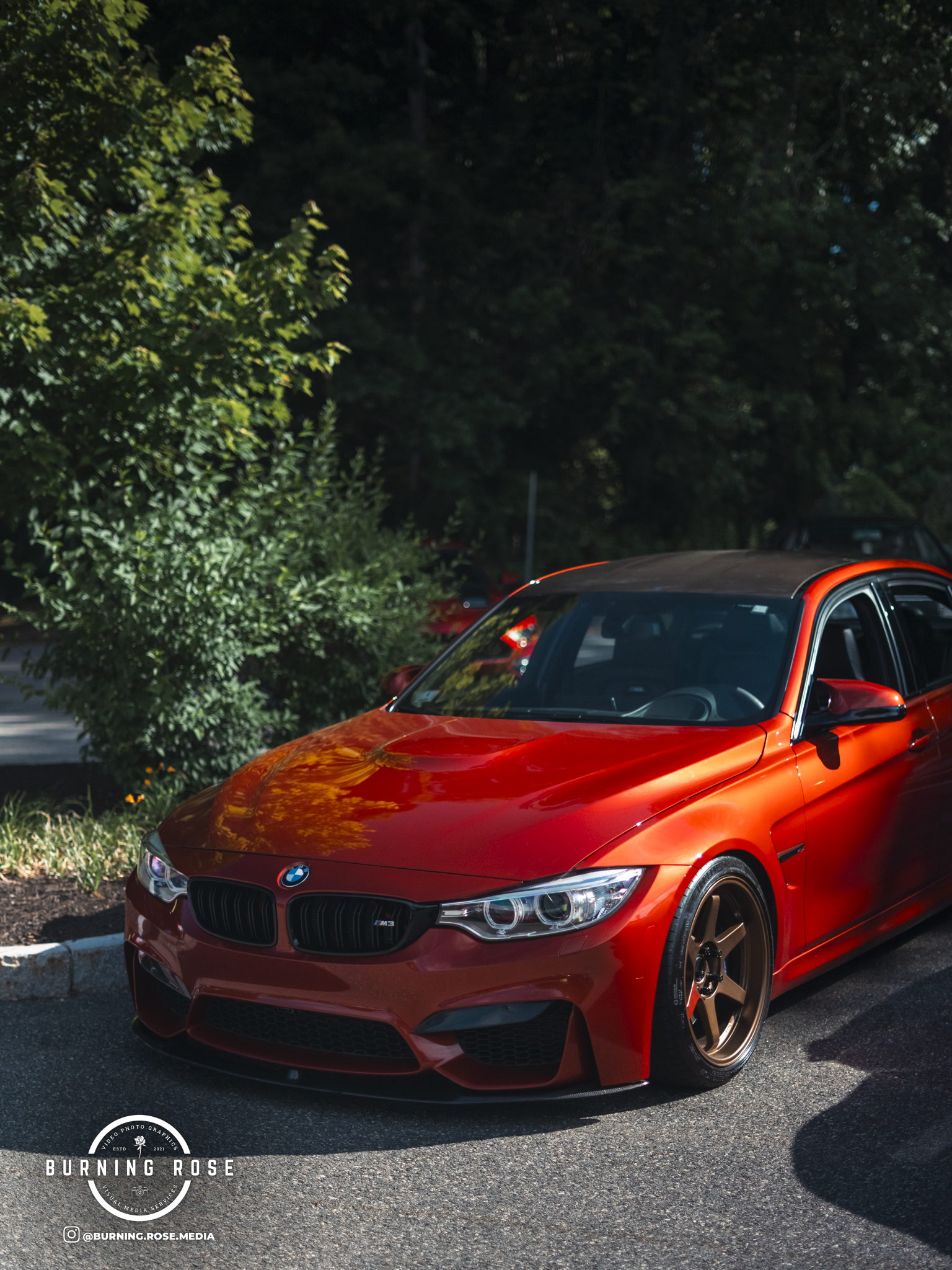 A bright orange BMW M3 sports car parked outdoors on a paved lot near green bushes and trees, with a nighttime setting.